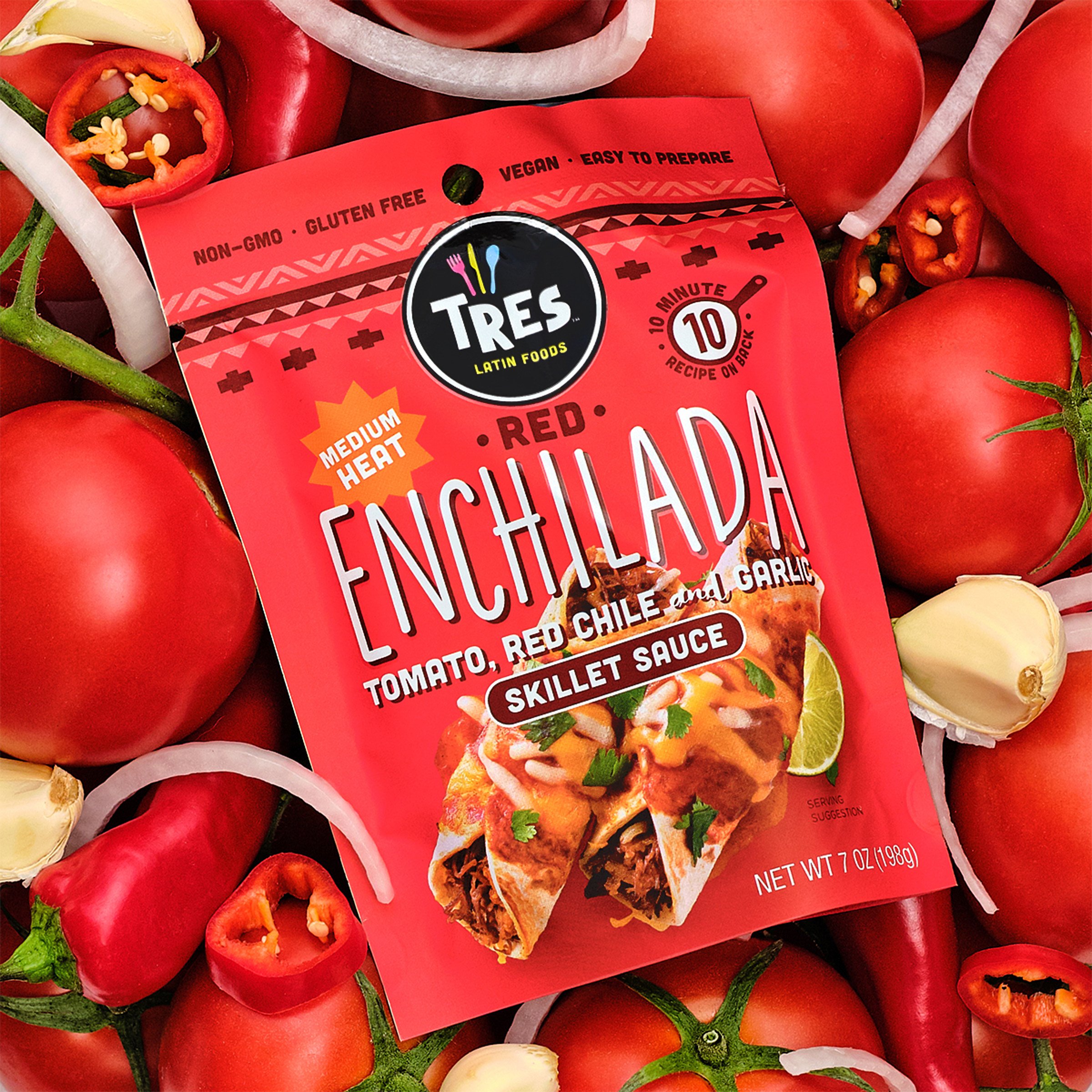 Close-up of a package of Tres Latin Foods enchiladas surrounded by fresh red tomatoes, sliced red chili peppers, garlic, and onions.
