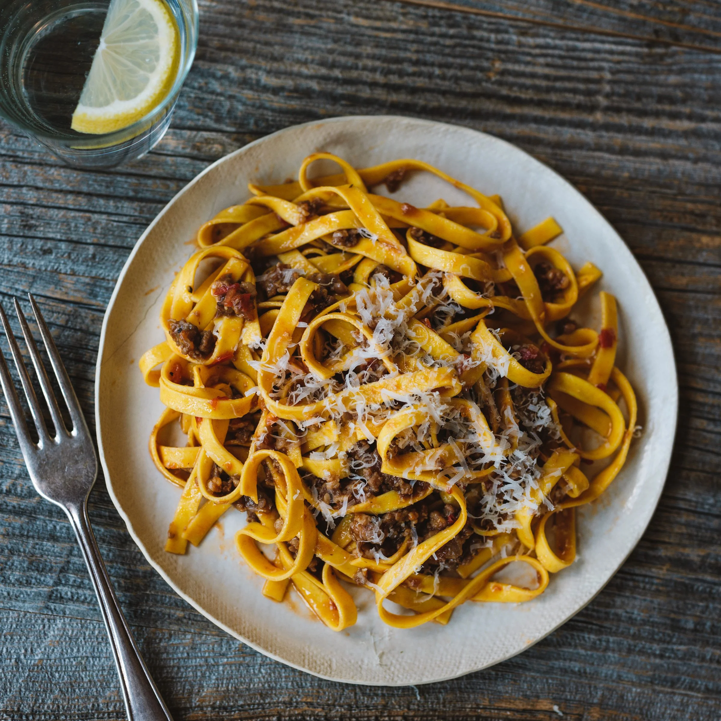 Plate of pasta with meat sauce and shredded cheese, garnished with a lemon slice, on a rustic wooden table with a glass of water with lemon slices.