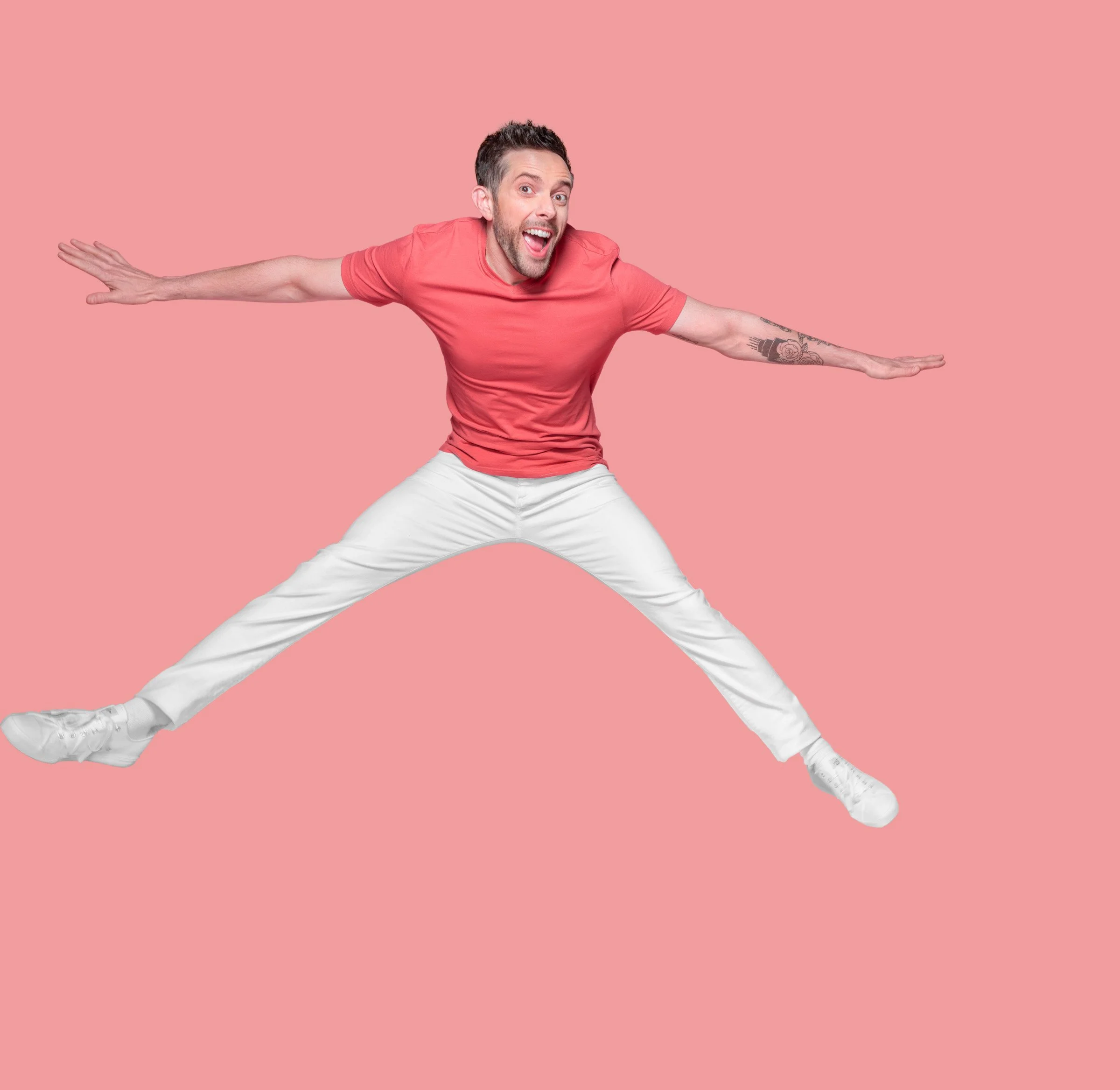 A man in a pink T-shirt and white pants doing a split with arms extended sideways against a pink background.