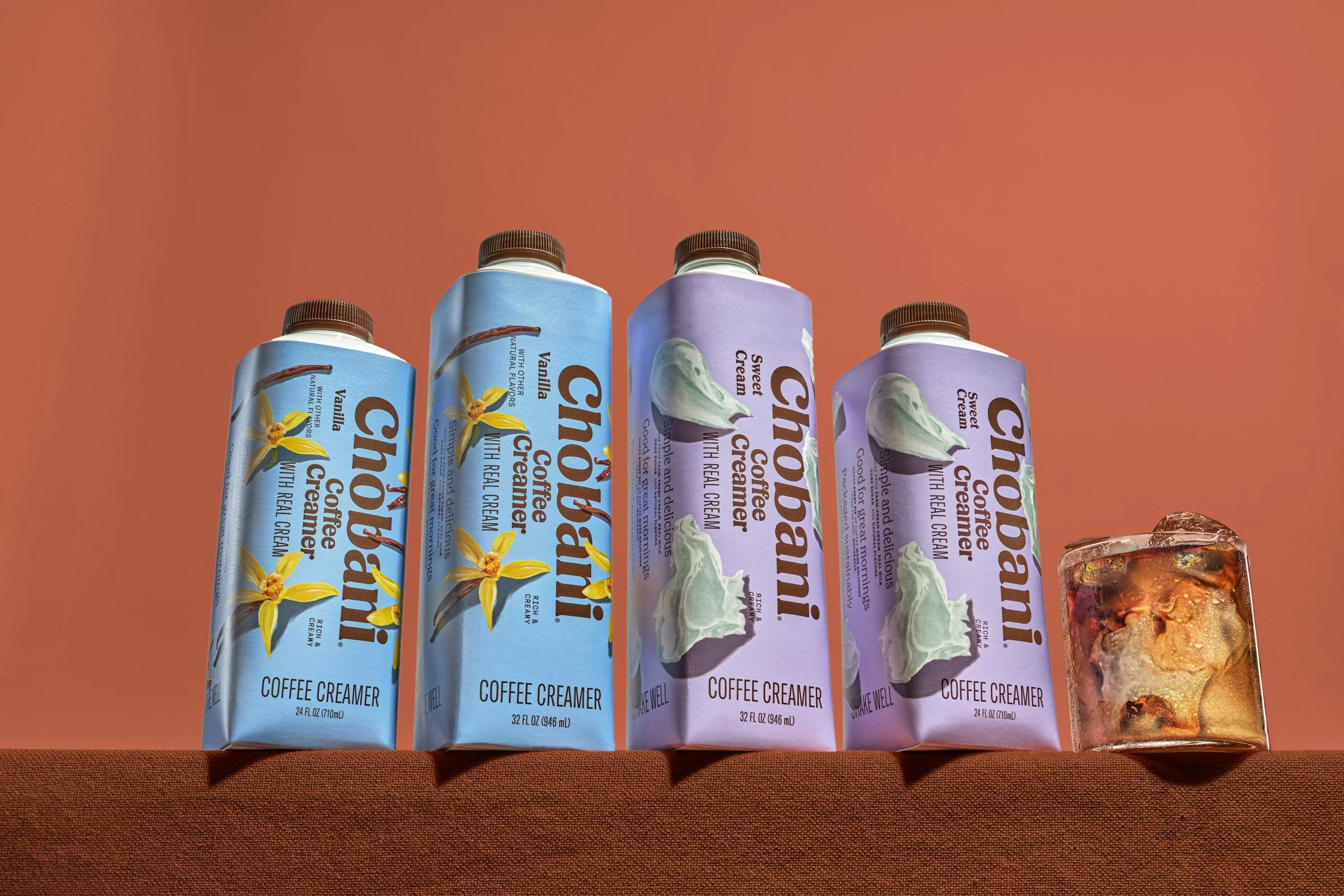Four cartons of Chobani coffee creamer in different flavors, placed on a surface with a reddish-brown background; partly visible glass of iced beverage on the right.