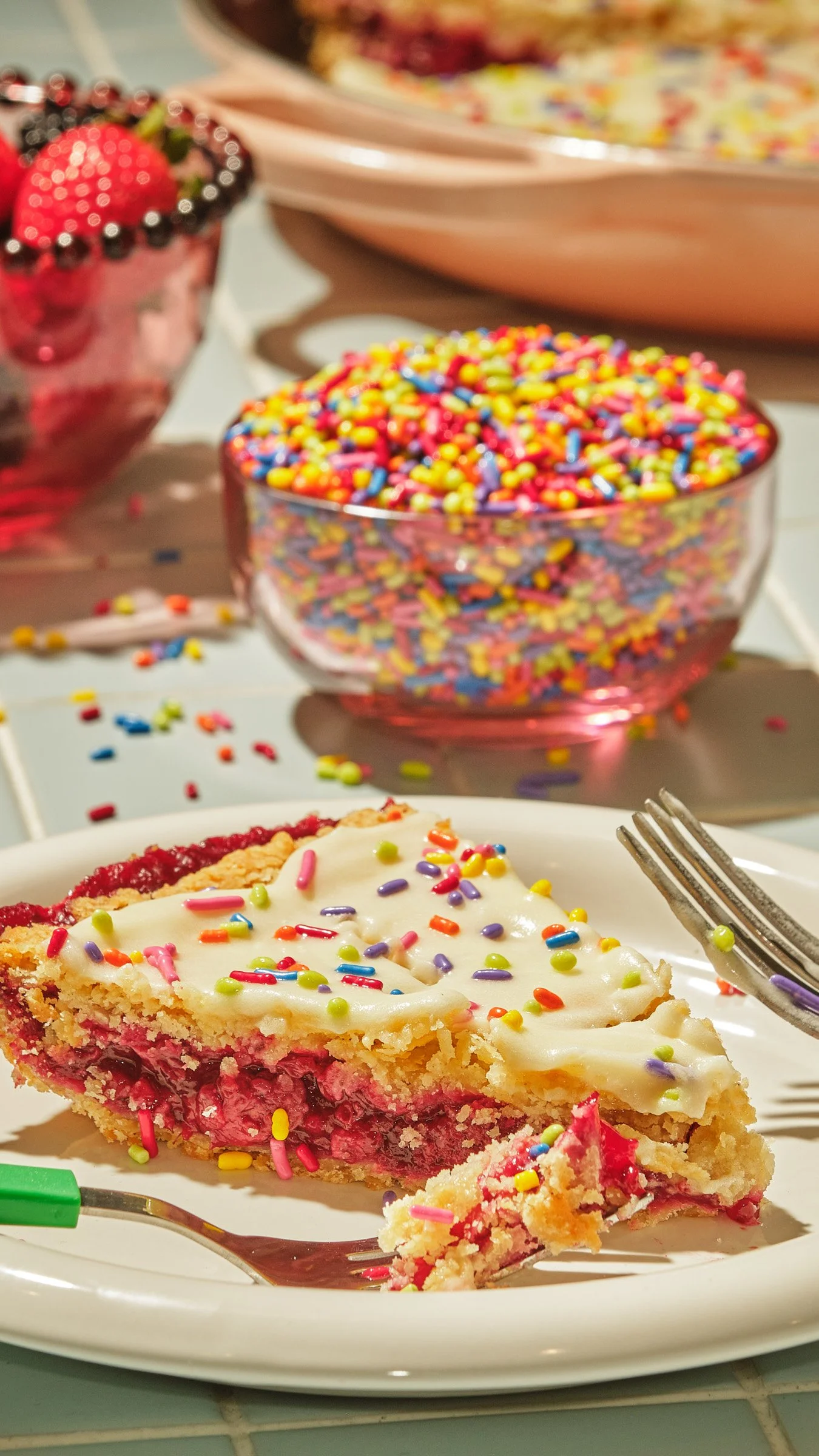 A slice of cherry pie with white icing and colorful sprinkles on a white plate, with a fork, in front of a bowl of rainbow sprinkles and a glass of berry dessert decorated with strawberries and blackberries.