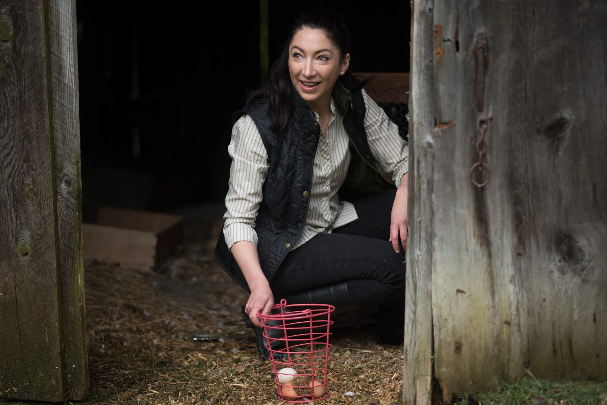 A woman crouches outdoors next to a wooden structure, holding a pink basket with eggs, surrounded by dirt and natural elements.