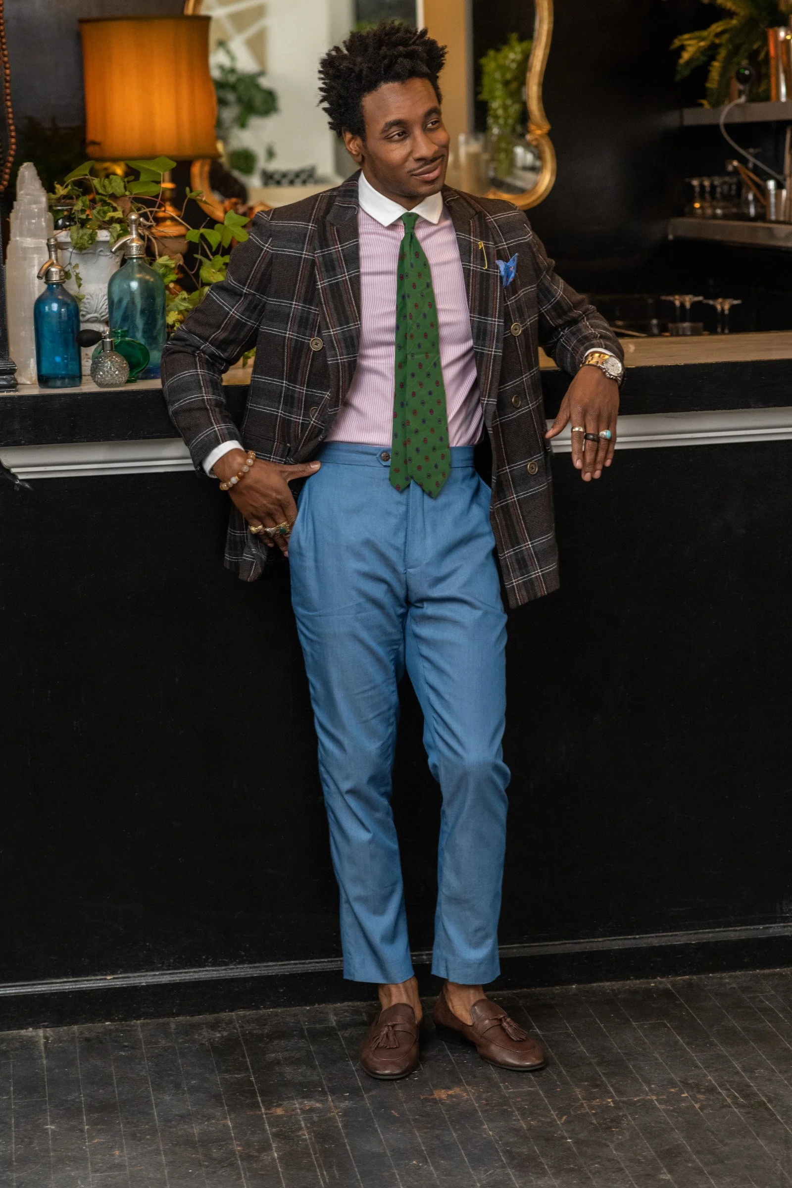 Man wearing a brown and black plaid blazer, pink and white striped shirt, green polka dot tie, blue trousers, and brown loafers, leaning on a kitchen counter with decorative bottles and plants in the background.