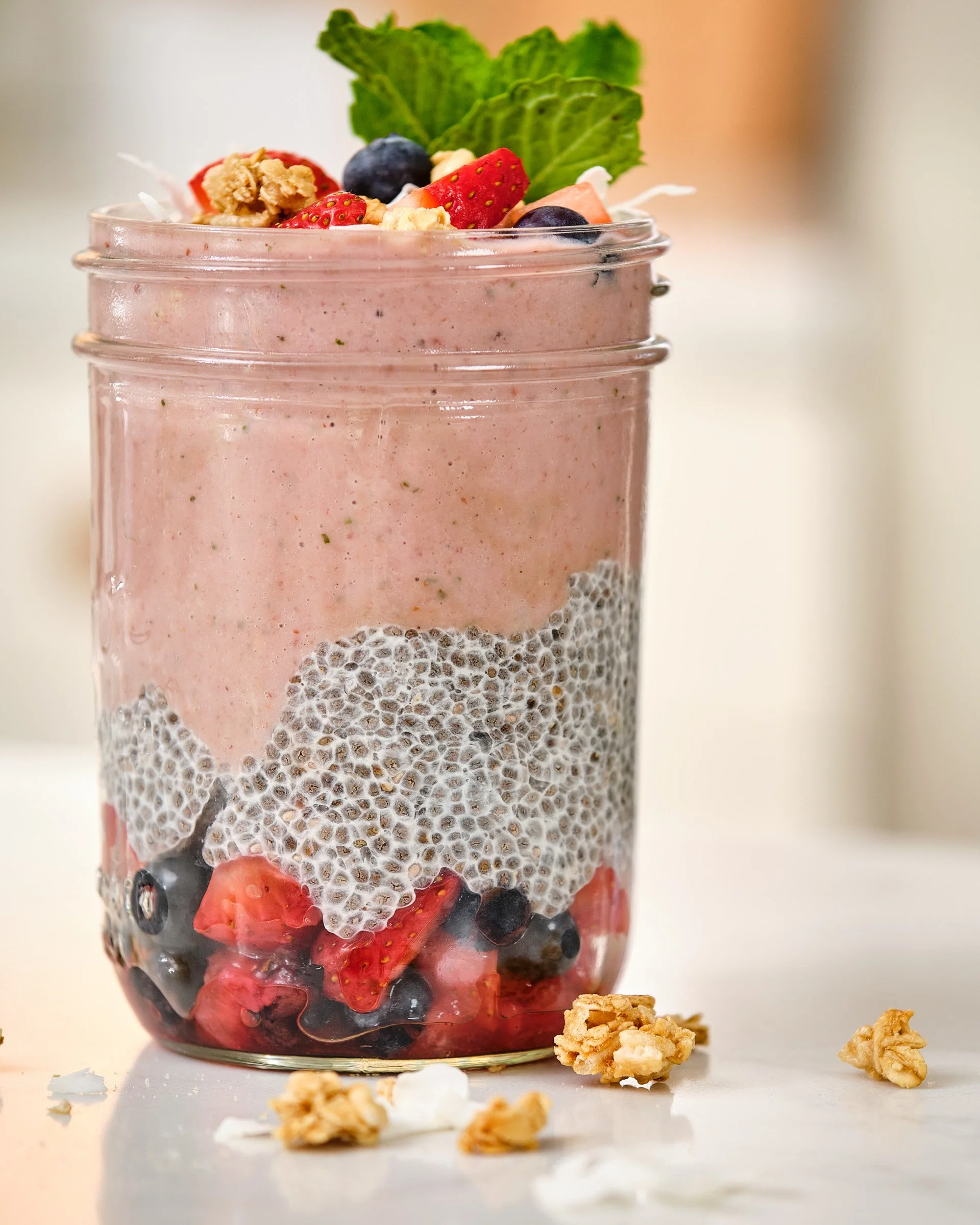 Close-up of layered berry smoothie in a glass jar, topped with strawberries, blueberries, granola, and mint leaves, with scattered granola and berry pieces on a white surface.