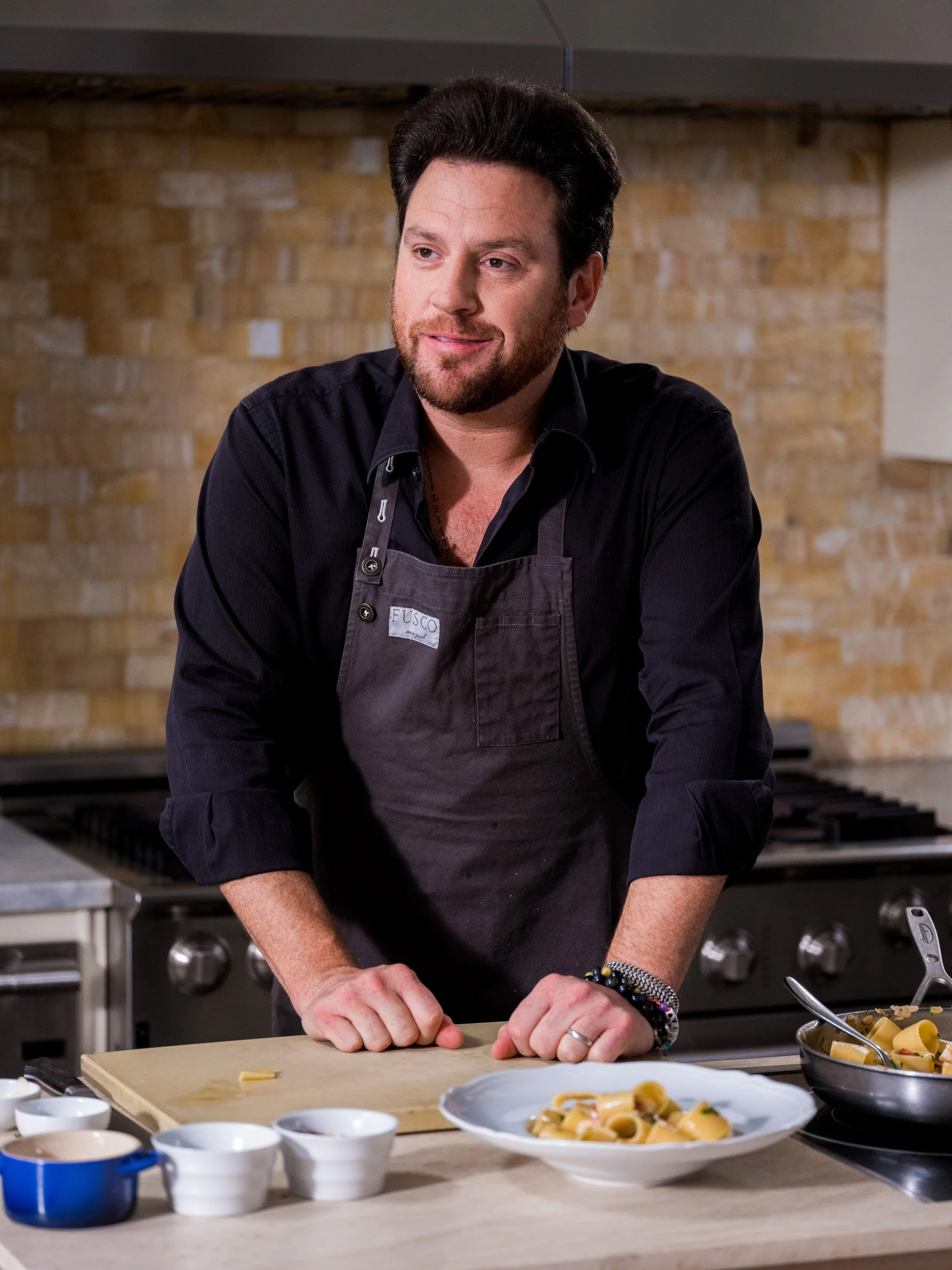 A man with dark hair, beard, and wearing a black shirt and apron preparing food in a kitchen with a wooden countertop and a brick wall in the background.