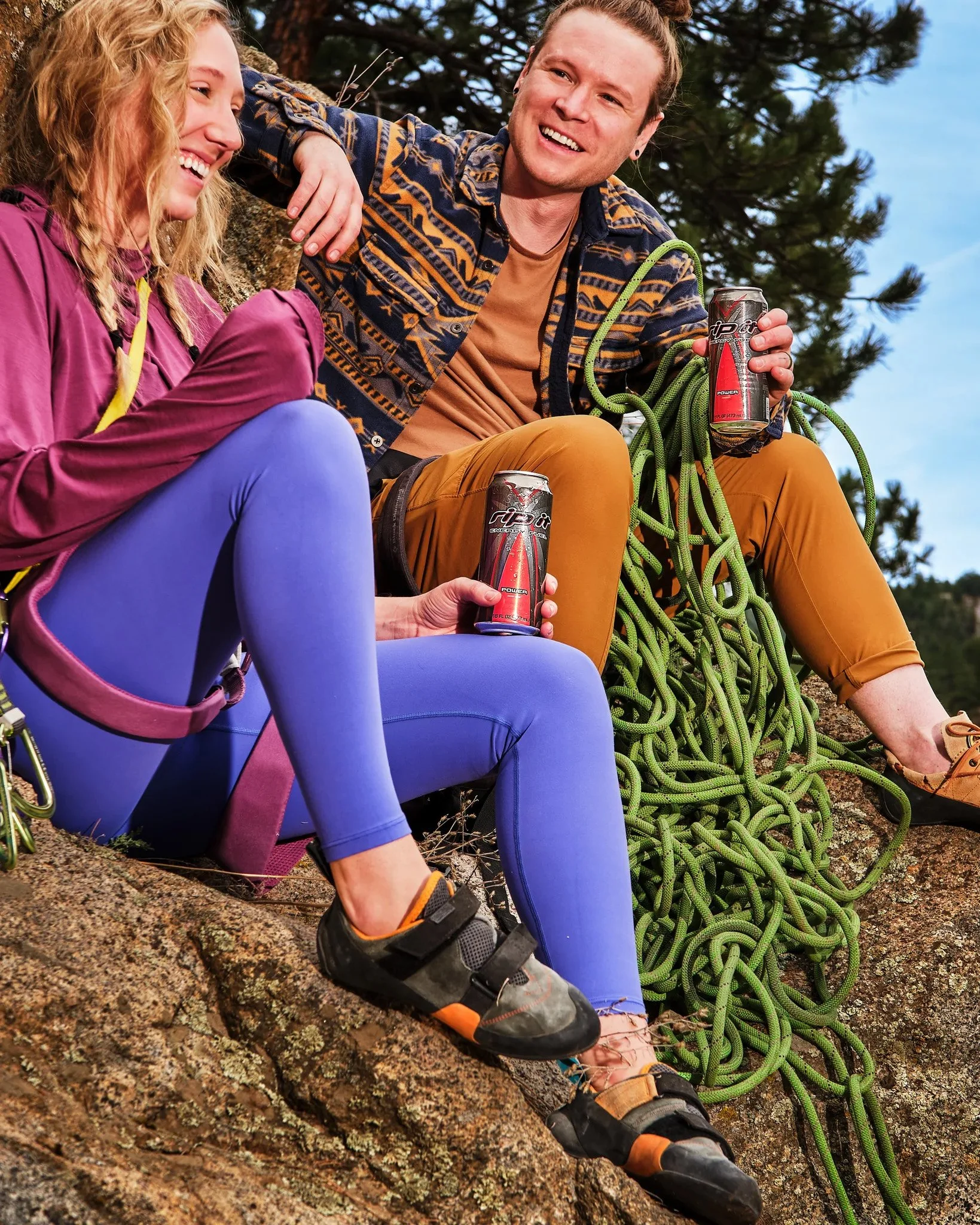 Two friends smiling and sitting on a rock during a mountain climbing trip, holding energy drink cans, with climbing gear and ropes nearby, surrounded by trees and clear sky.
