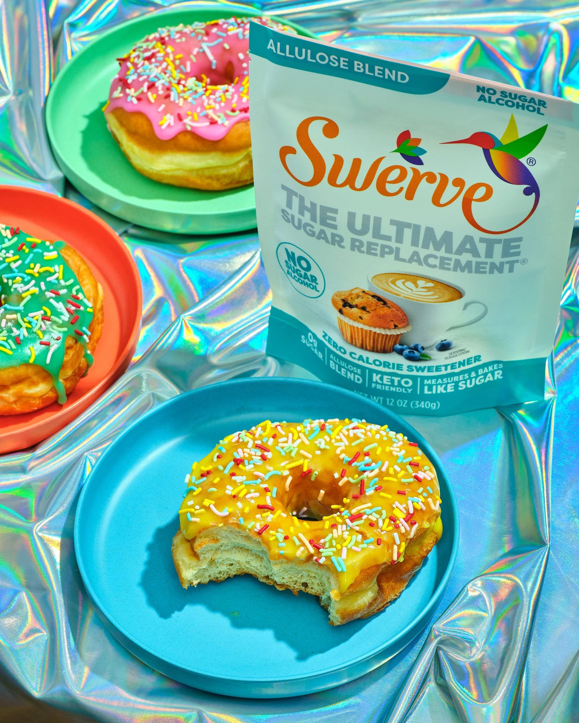 A donut with pink icing and colorful sprinkles on a green plate beside another donut with yellow icing and sprinkles on a blue plate, with a package of Swerve allulose sweetener in the background. The background features iridescent, holographic mater