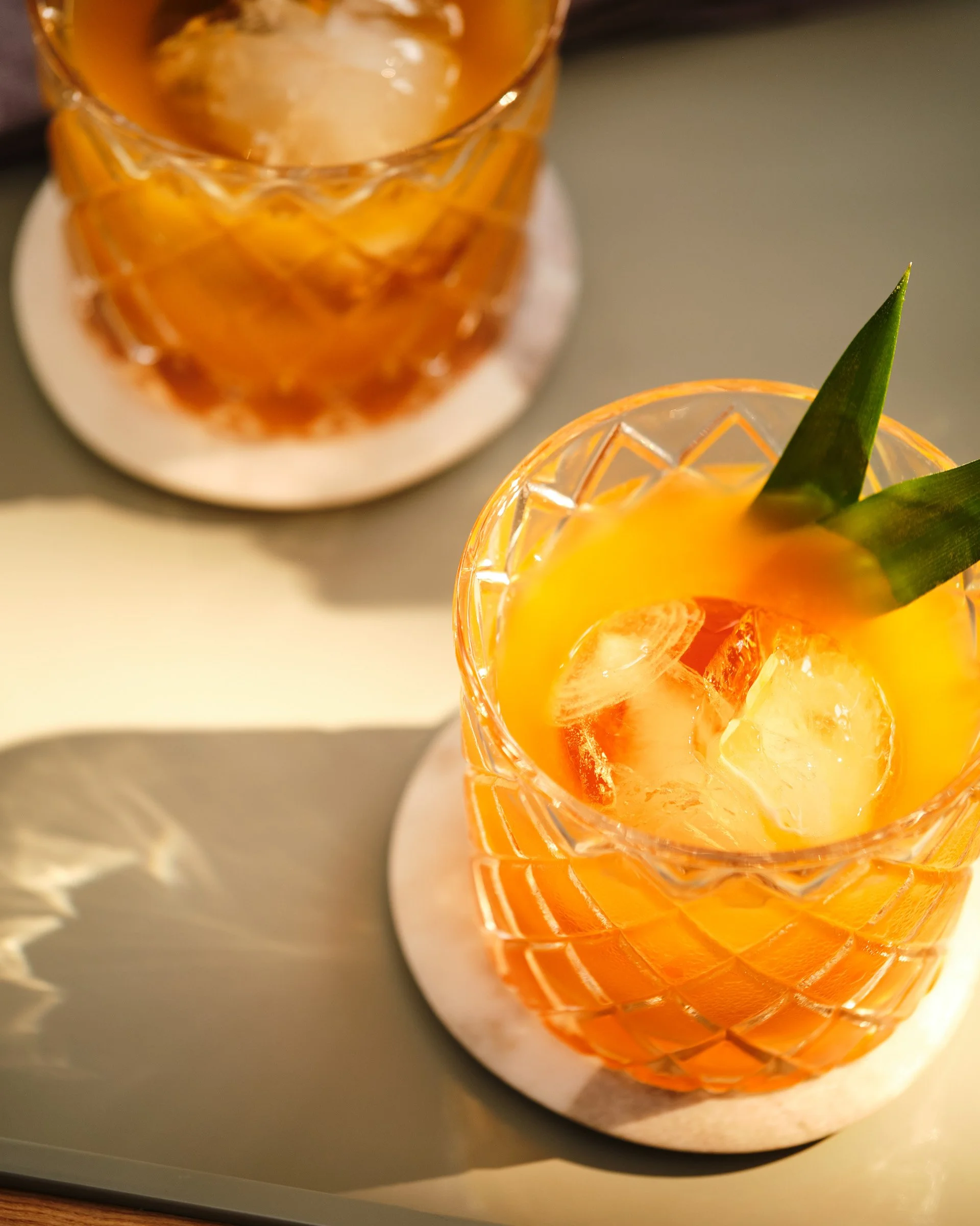 Close-up of a glass filled with an orange-colored drink, garnished with pineapple slices and green pineapple leaves, on a white coaster.