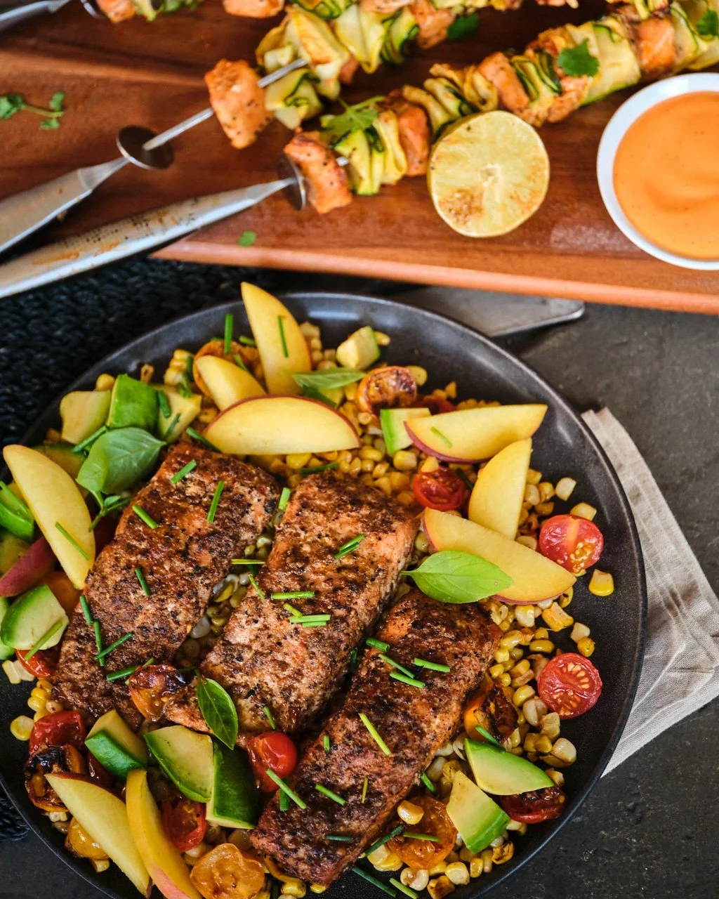 Grilled fish fillets on a bed of corn, cherry tomatoes, sliced pears, zucchini, and herbs, garnished with green herbs, served in a black skillet.