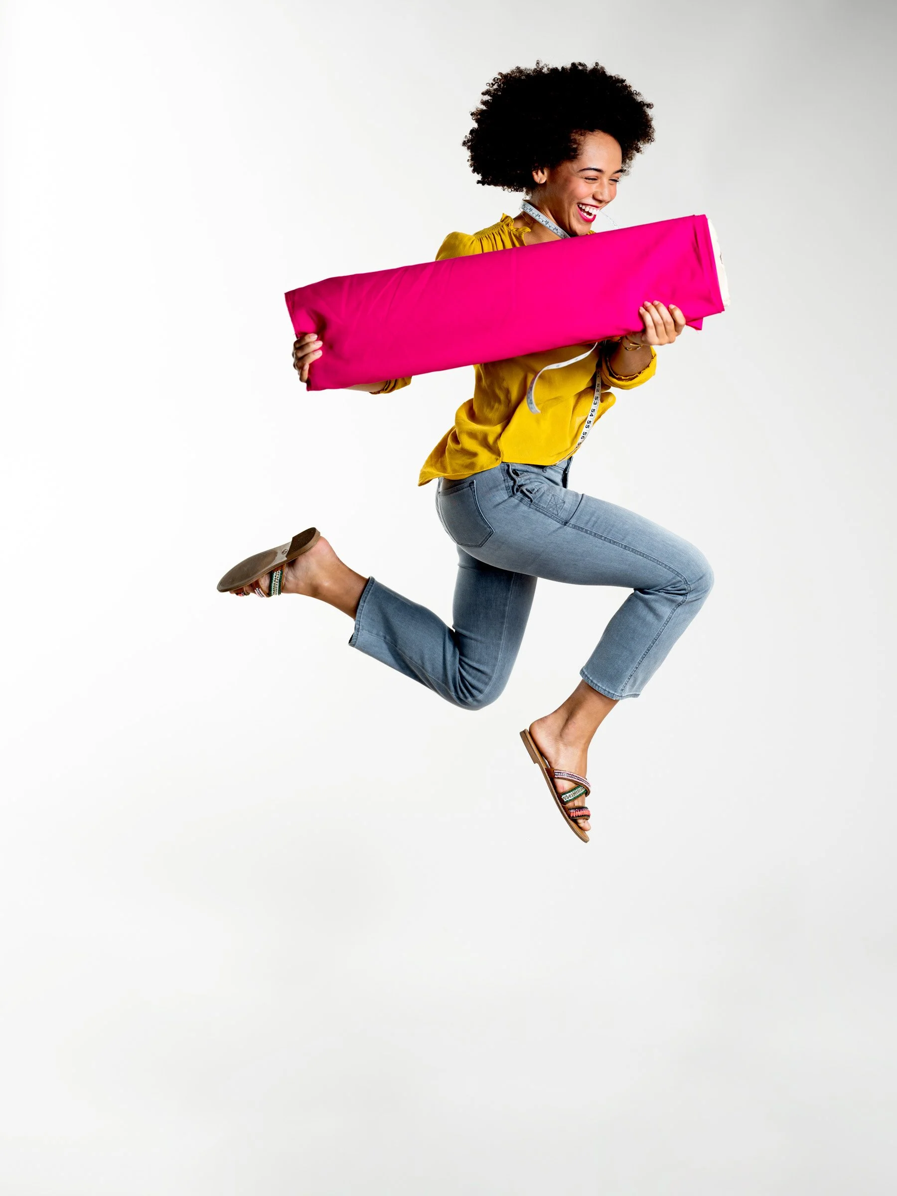 A woman with curly hair, wearing a yellow top, jeans, and sandals, is jumping while holding a large pink cloth or cushion and smiling.