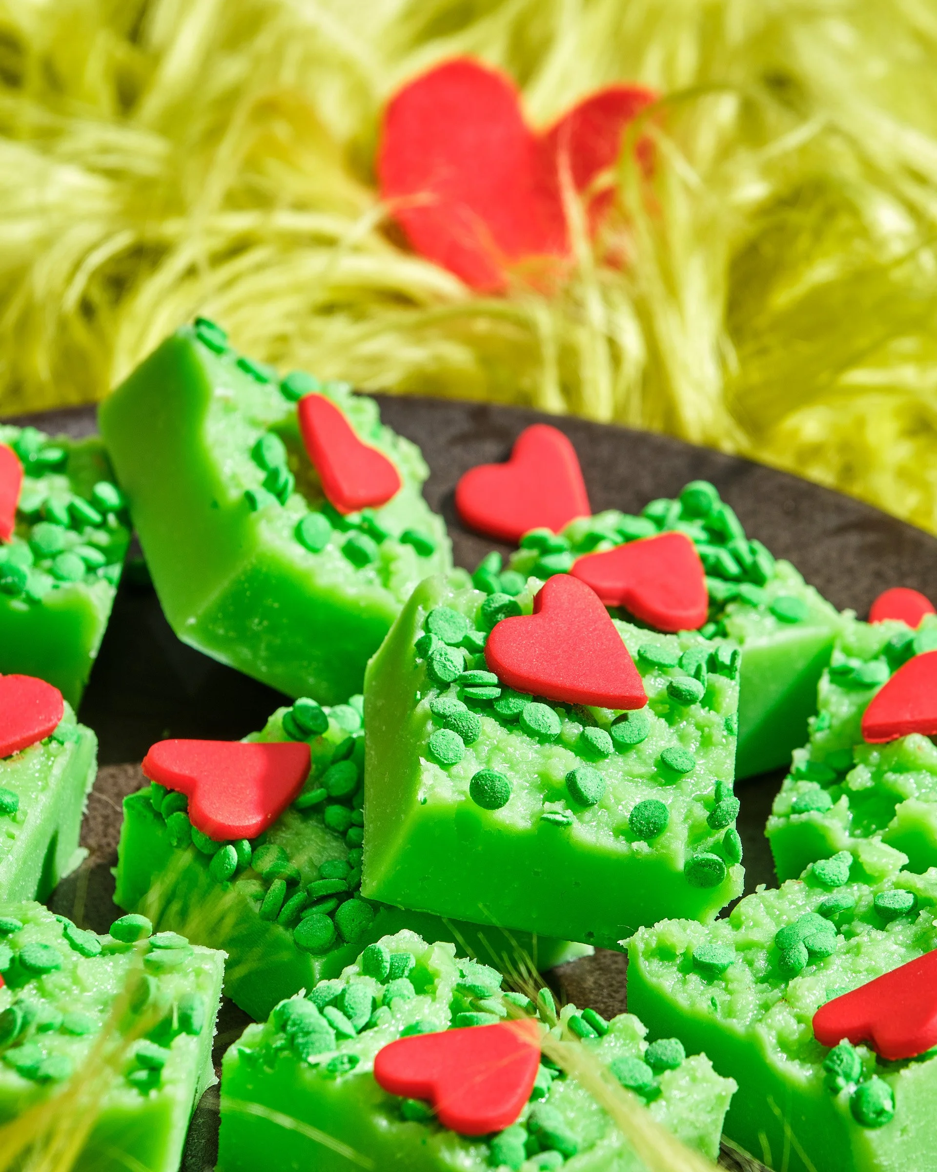 Close-up of green decoratively shaped soaps with red heart-shaped toppers and green sprinkles, with blurred yellow background.