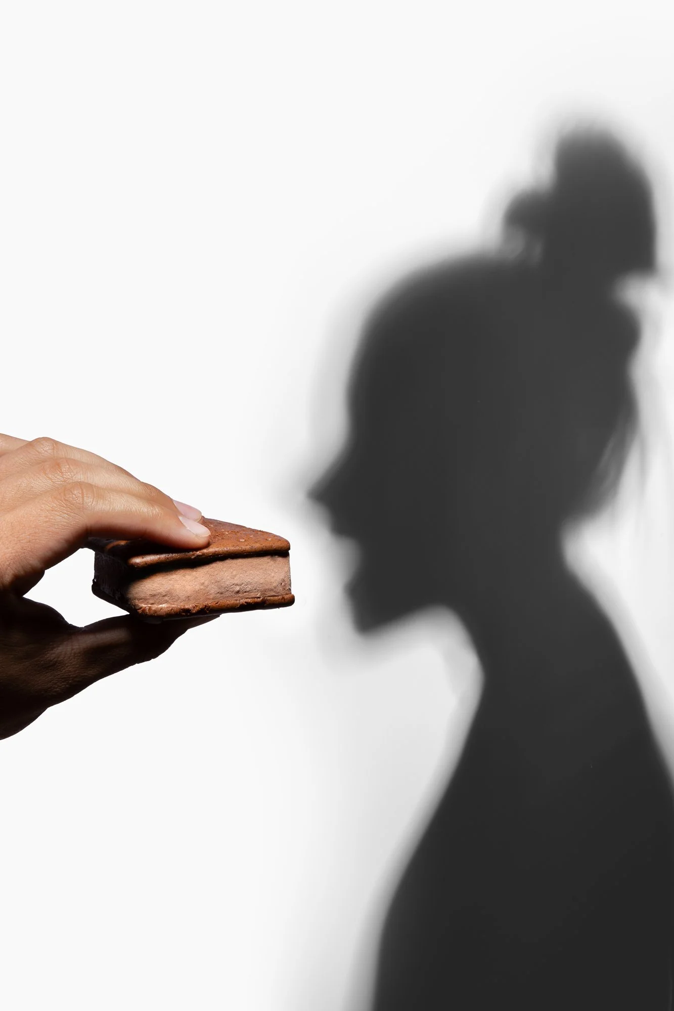 A hand holds a chocolate ice cream sandwich against a white background. The shadow of a person cast on the wall bites at sandwich.