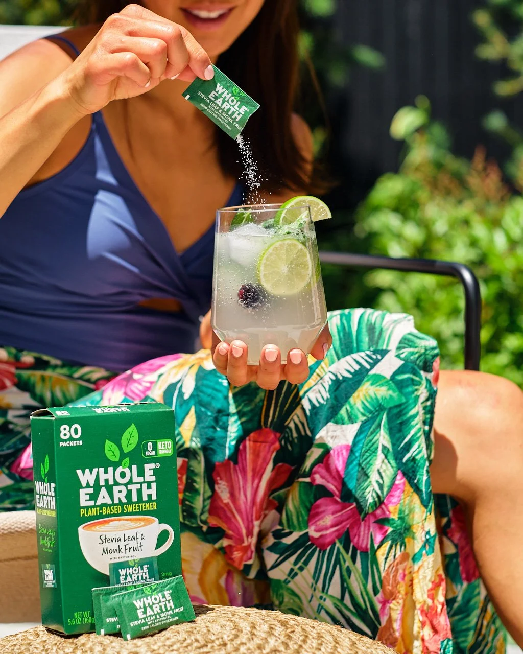 A woman in a blue top is watering a glass of lemonade with lime slices and blackberries, sugar packets are sprinkled into the drink, and a box of Whole Earth plant-based sweetener is on the table in front of her. She is wearing colorful tropical patt