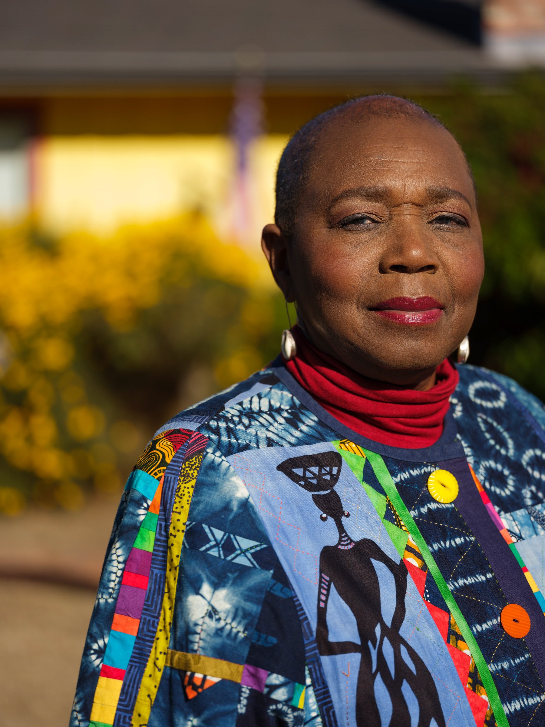 Close-up of an elderly African American woman with short hair, wearing a colorful, patterned jacket with buttons and a red turtleneck, standing outdoors with blurred greenery in the background.