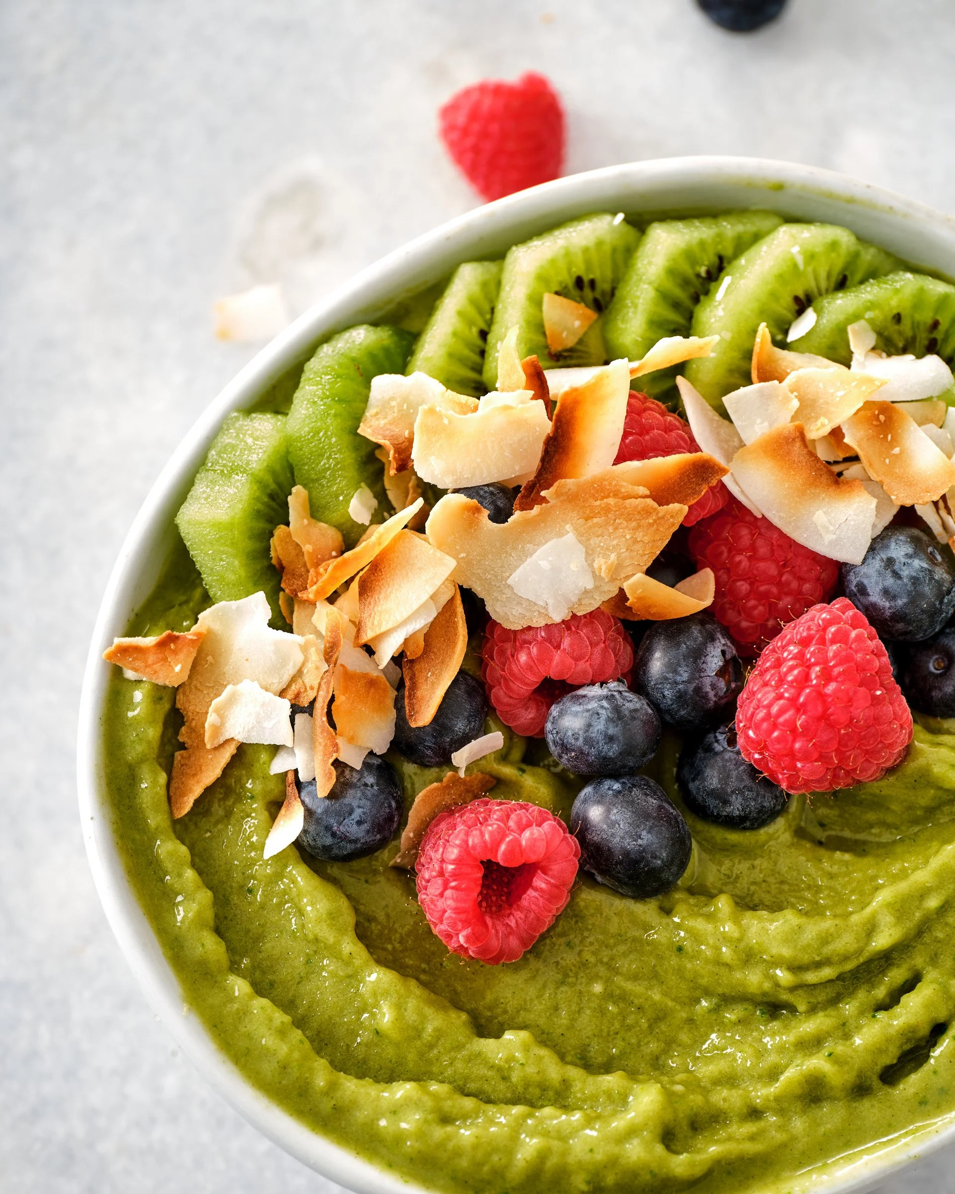Top view of a smoothie bowl with sliced kiwi, raspberries, blueberries, shredded coconut, and coconut flakes in a white bowl on a gray surface.