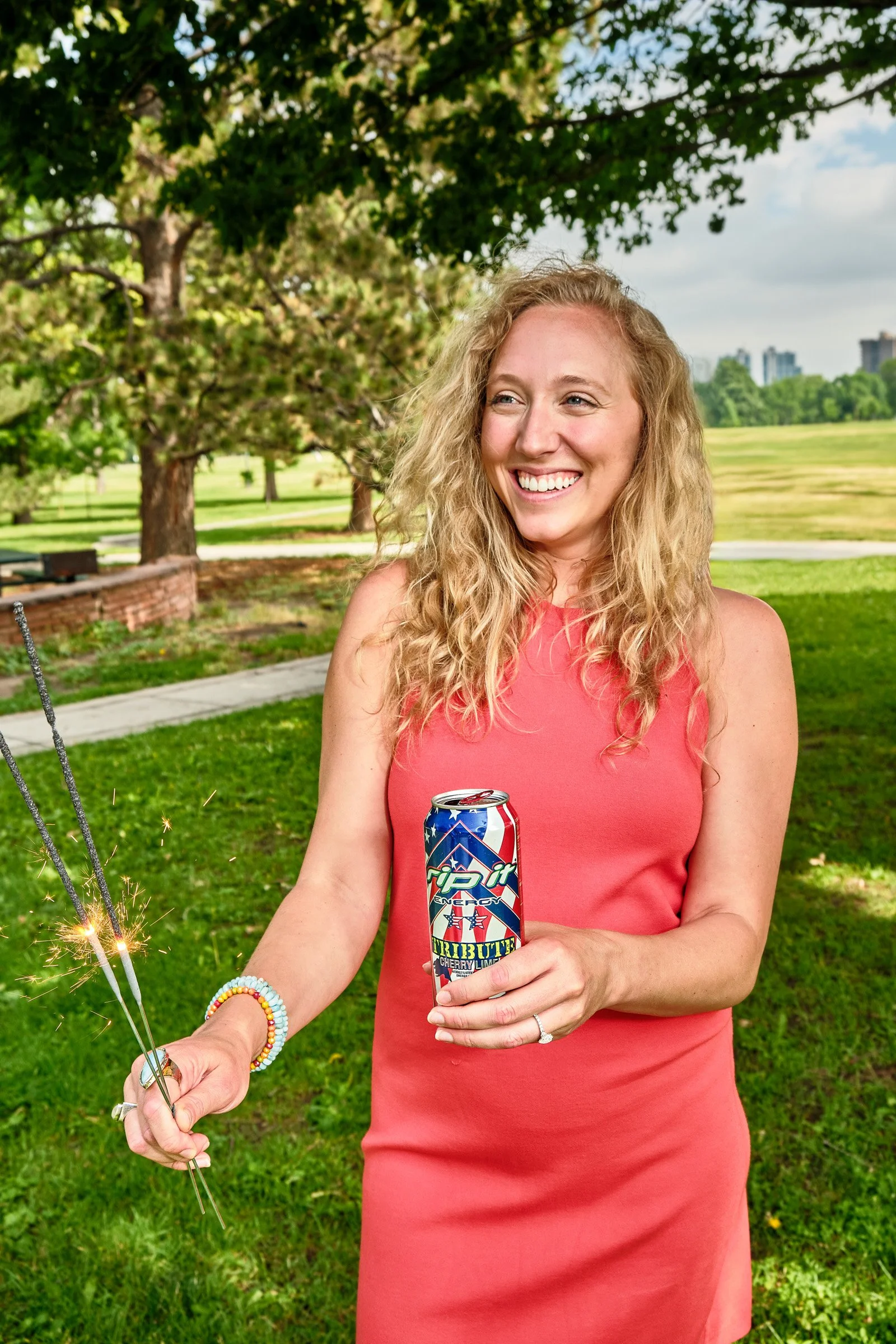 A smiling woman with curly blonde hair in a pink dress holding a can of 7UP Plus Cherry Lime in a park with trees and green grass, celebrating with sparklers.
