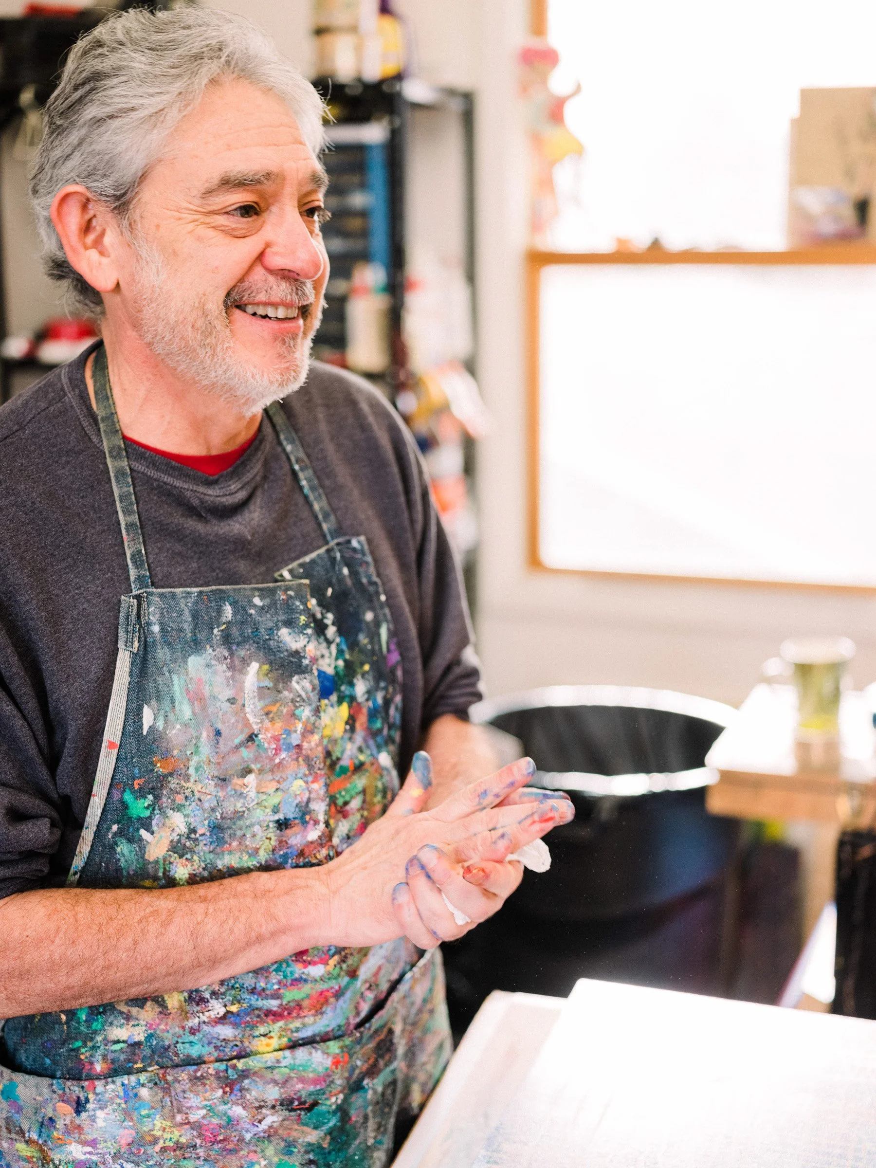Older man with gray hair and beard wearing a paint-splattered apron, smiling in an art studio or classroom with art supplies and a window in the background.