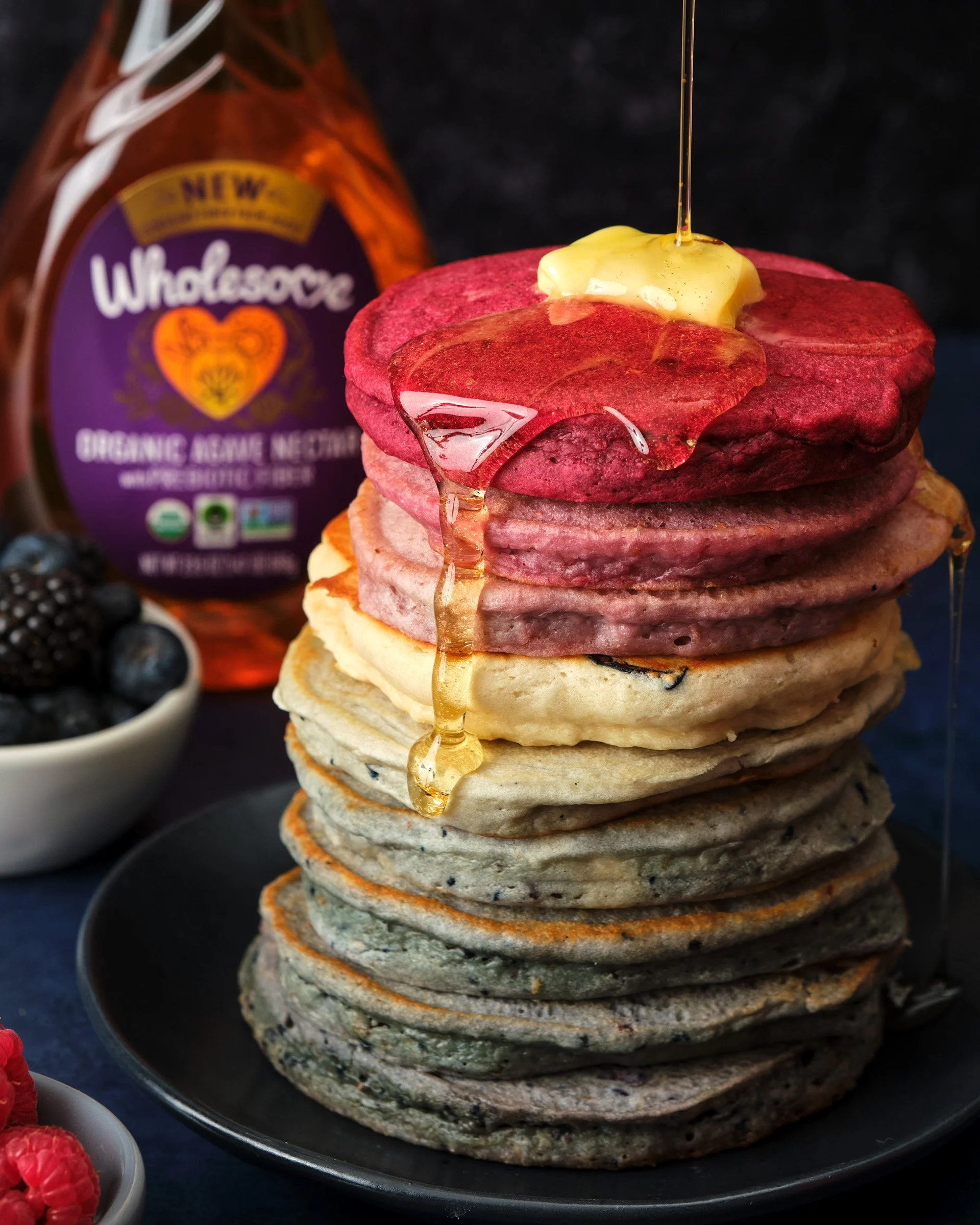 Stack of six pancakes topped with slices of beet and a spoonful of butter, with syrup being poured on top, on a dark plate with a small bowl of blueberries in the background and a bottle of organic almond milk.
