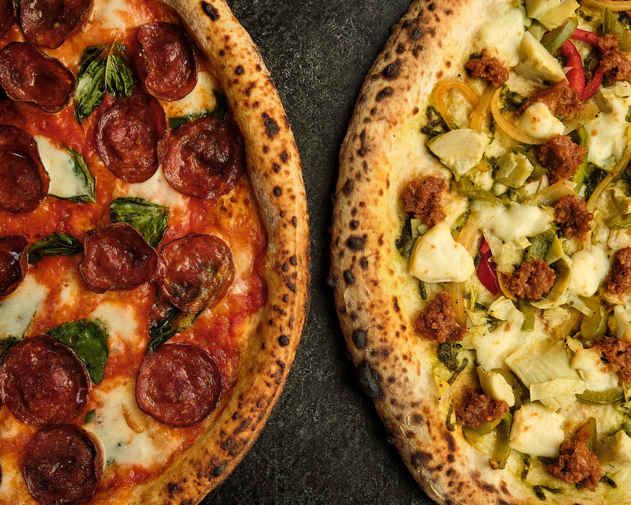 Close-up of two pizza slices on a dark surface. The left slice is topped with pepperoni, basil, and cheese. The right slice has a variety of vegetables including artichokes, green peppers, onions, and ground meat.