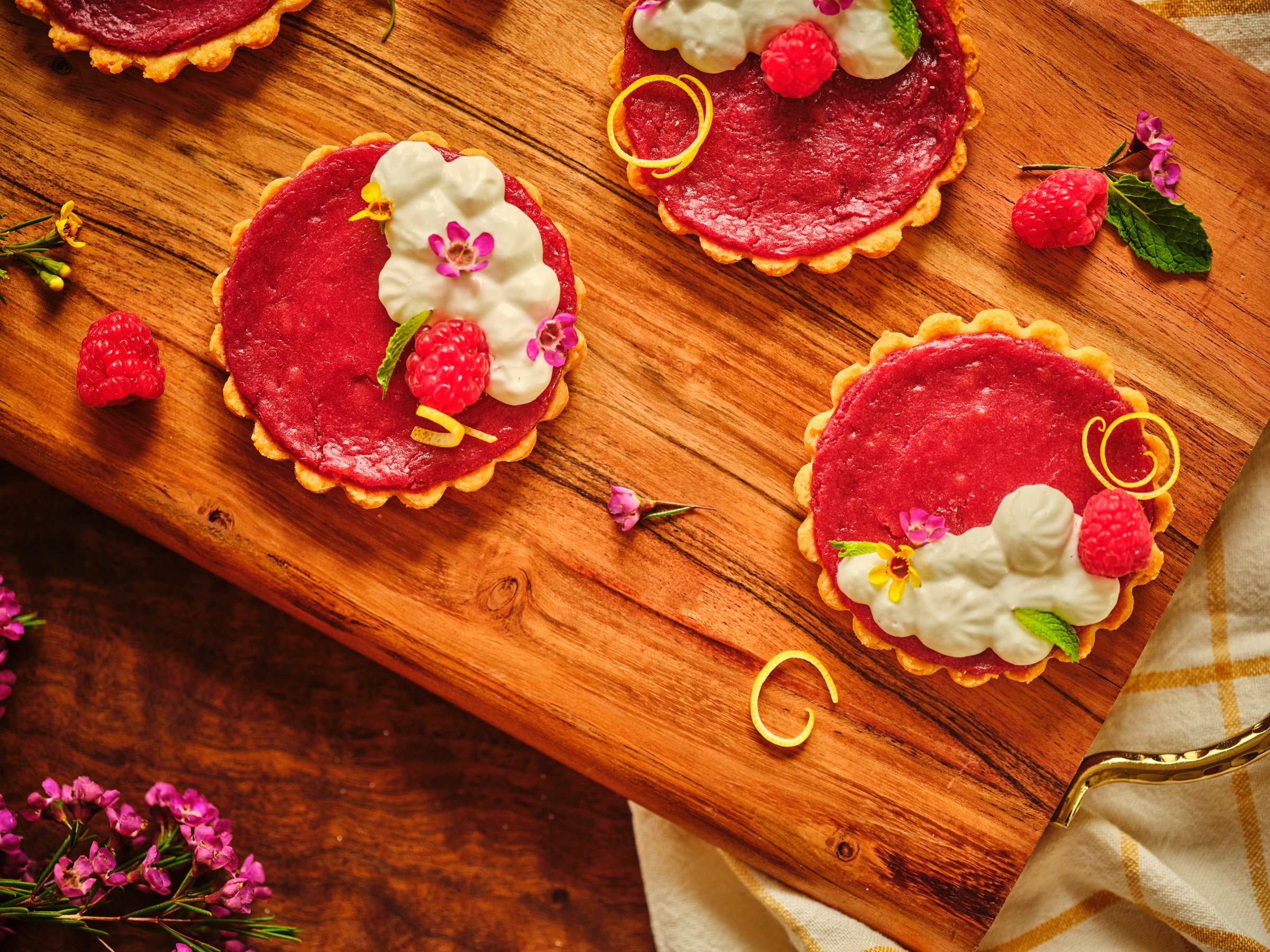 Multiple raspberry tartlets with decorative cream, edible flowers, and raspberry topping placed on a wooden surface with scattered raspberries, flowers, and a yellow-and-white checkered cloth.