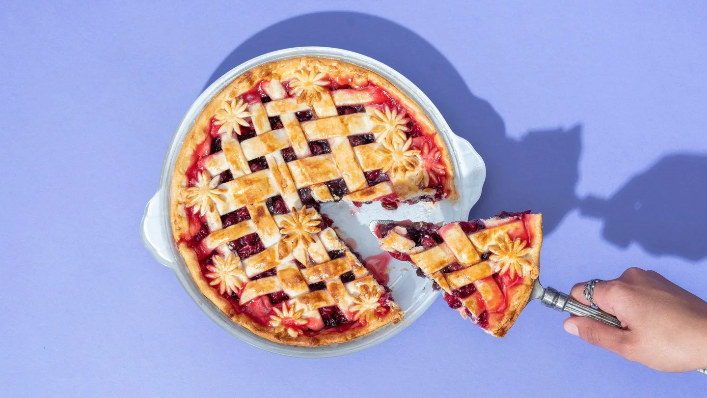 A hand is holding a pie server with a slice of cherry pie, near a whole cherry pie in a glass dish on a purple background.