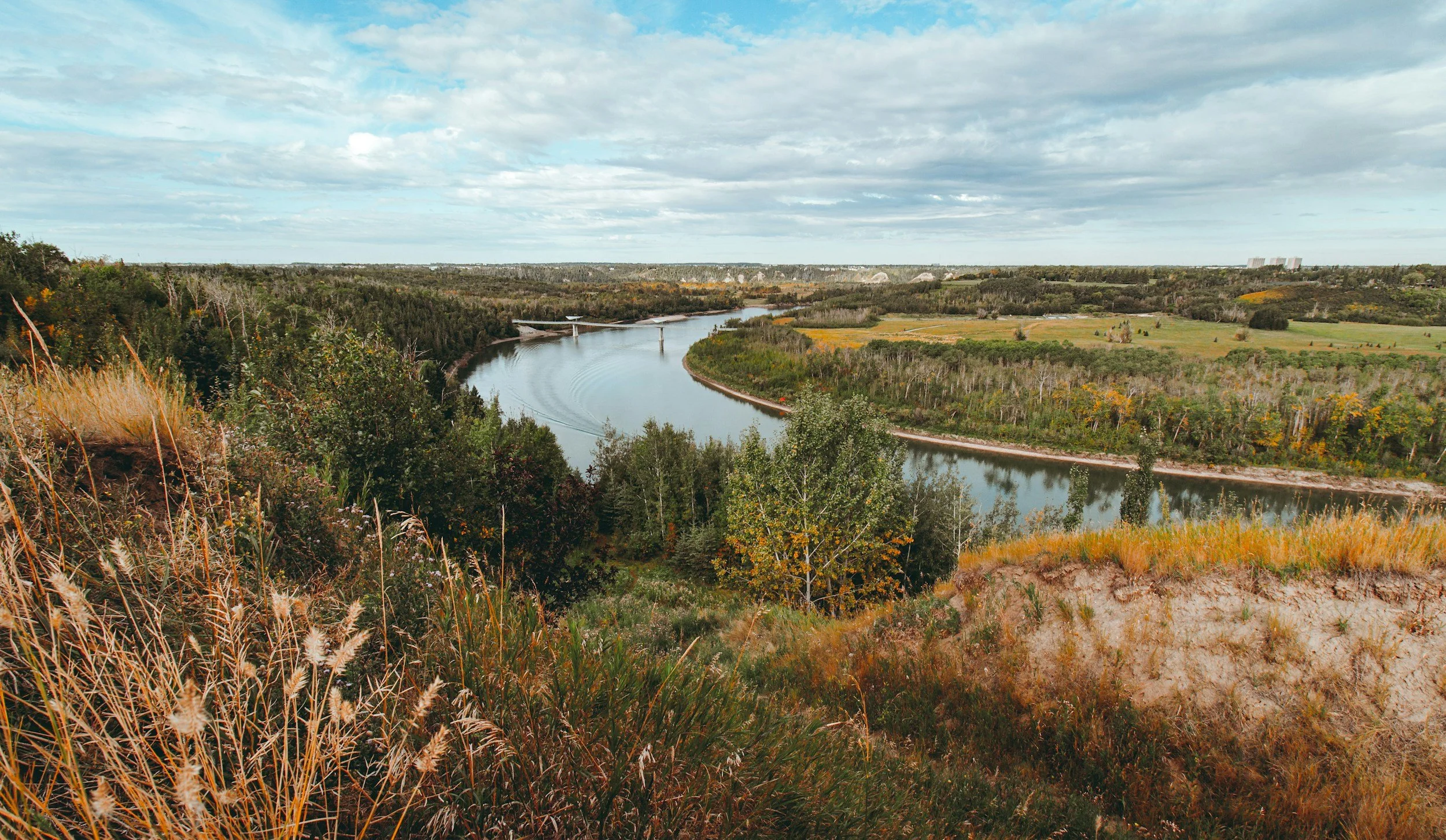 South Saskatchewan River  winding through a landscape with grassy hills and trees, and an arch bridge crossing the river in the distance under a partly cloudy sky.