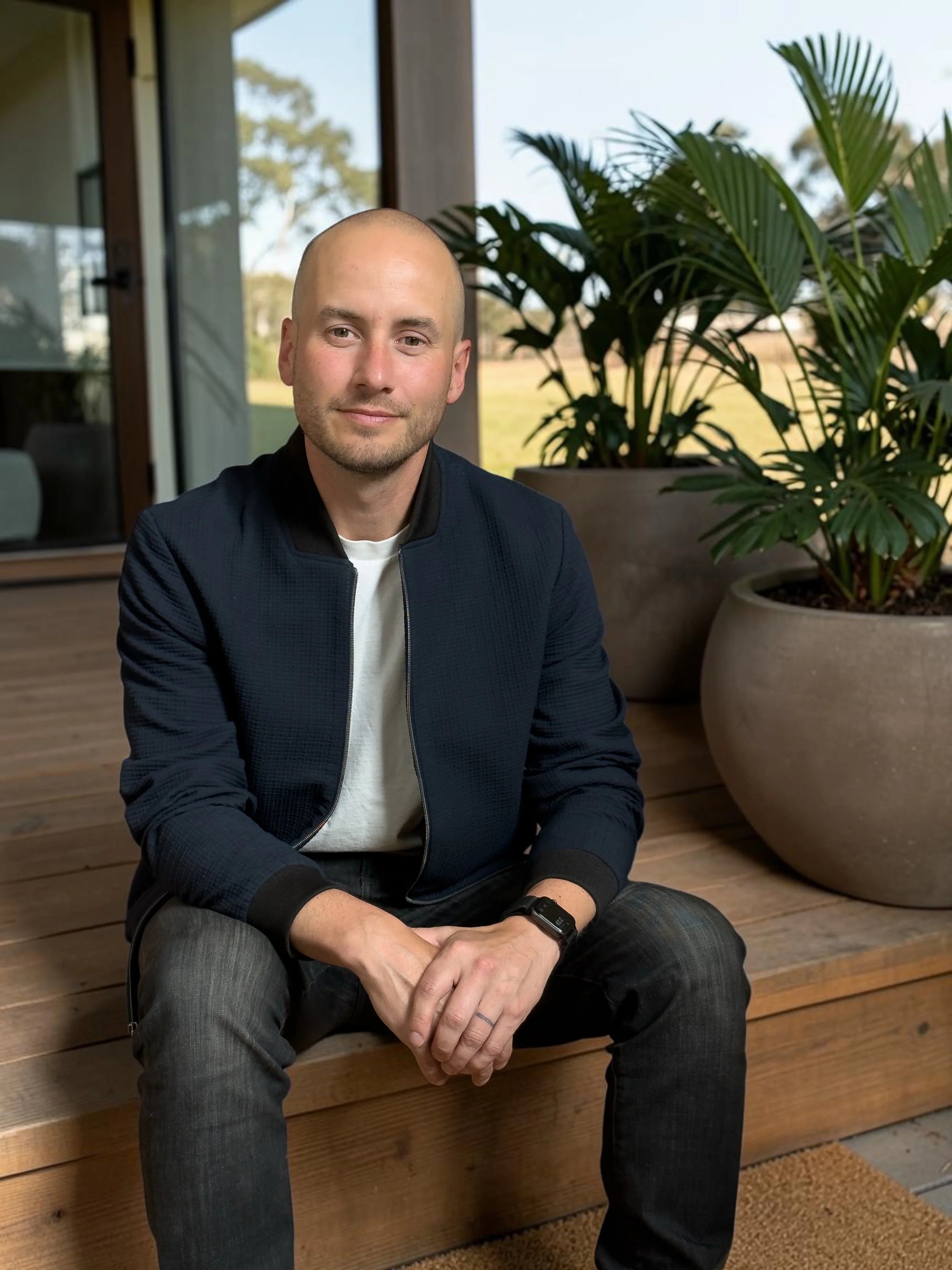 A man with a shaved head and beard sitting outdoors on a wooden deck, wearing a black jacket, white shirt, and jeans, with large potted plants and a house in the background.