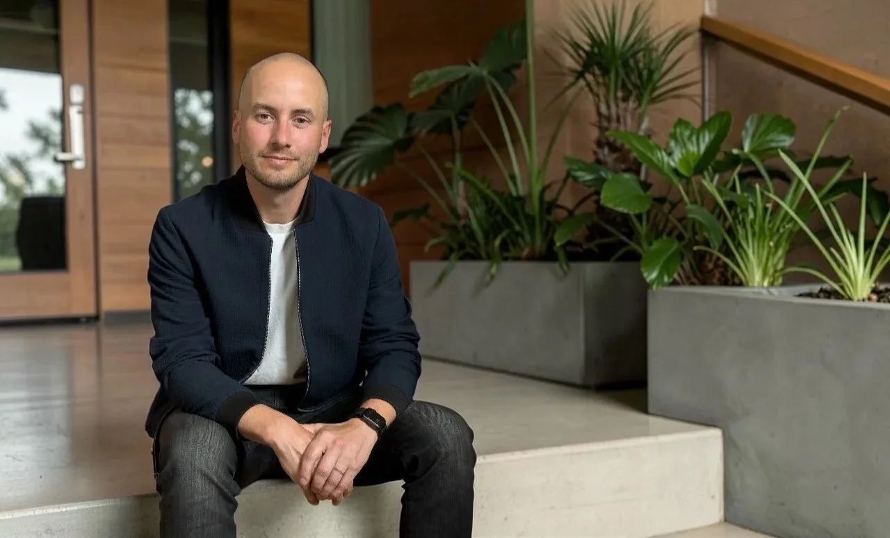 Mark Neudorf sitting on stairs indoors, with greenery in large planters beside him.