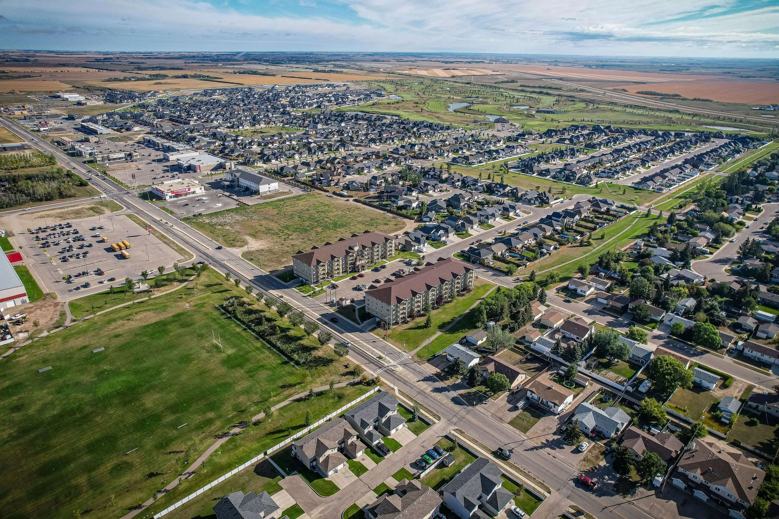 Aerial view of City of Warman with residential houses, apartment buildings, parking lots, and open green spaces, with fields and farmland in the distant background under a partly cloudy sky.