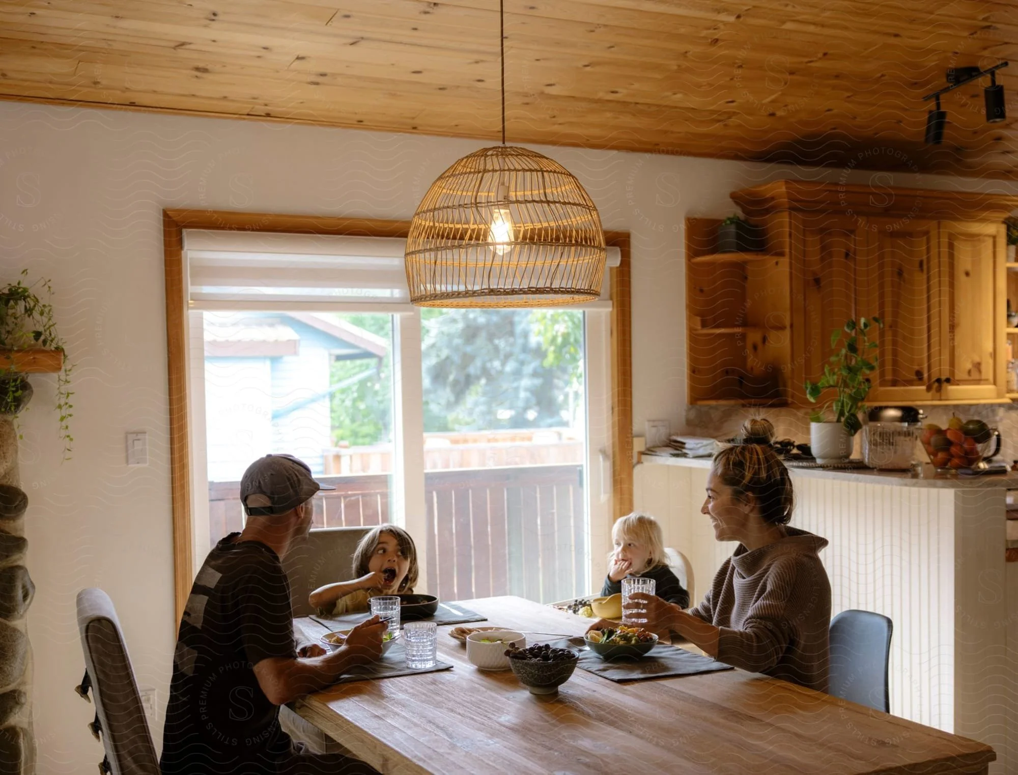 Family gathered around a dining table with food in a bright kitchen, with a wooden ceiling, window, and hanging light fixture.