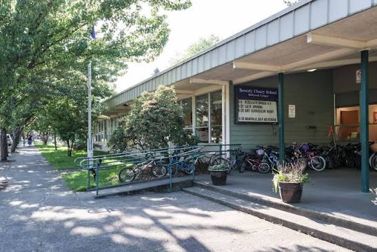 The side of a building with a ramp and bicycle parking, next to a sidewalk with trees and grass.