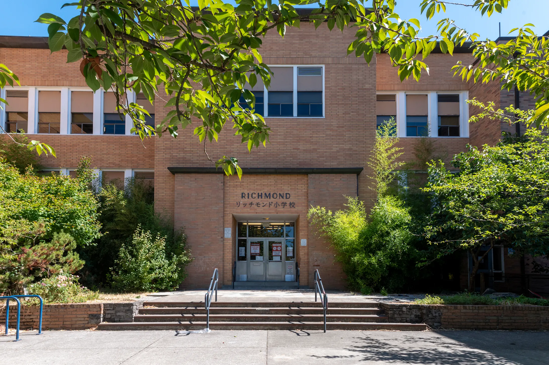 The front entrance of Richmond Elementary School, a brick building with a sign in Japanese characters, surrounded by green trees with bright sunlight.