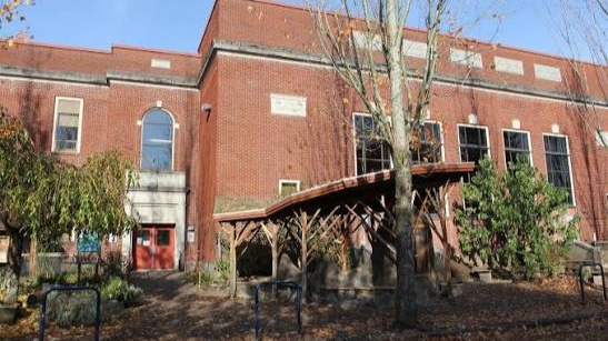 A red brick building with windows and a small covered shelter in front, surrounded by trees and bushes.