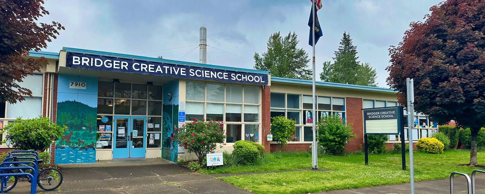 Exterior of Bridger Creative Science School with a blue sign, colorful mural on the building, and a well-maintained lawn with trees and bushes.