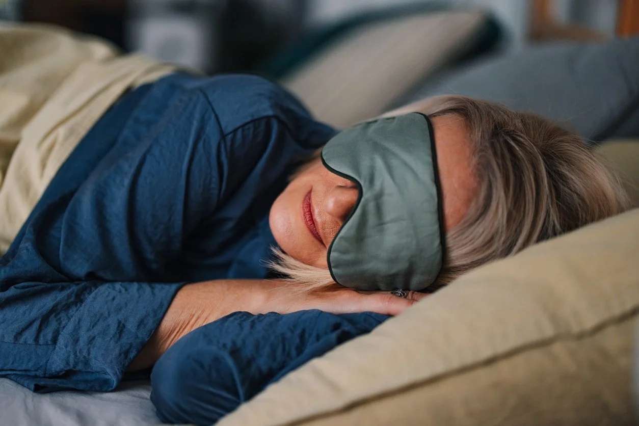 Older woman sleeping on her side in bed with a sleep mask over her eyes.
