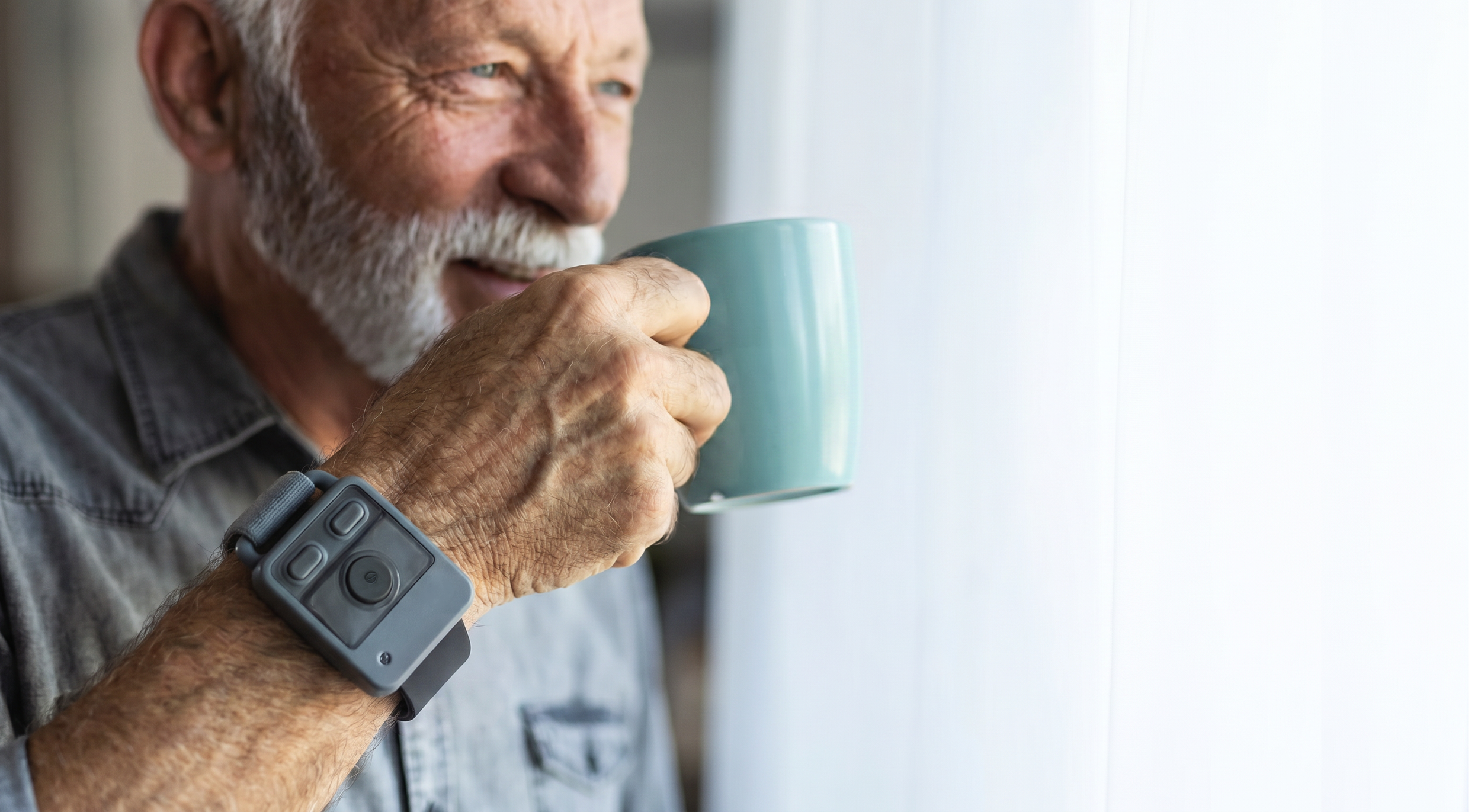 An elderly man with a white beard, wearing a gray shirt and a smartwatch, is drinking from a light blue mug while looking out a window.