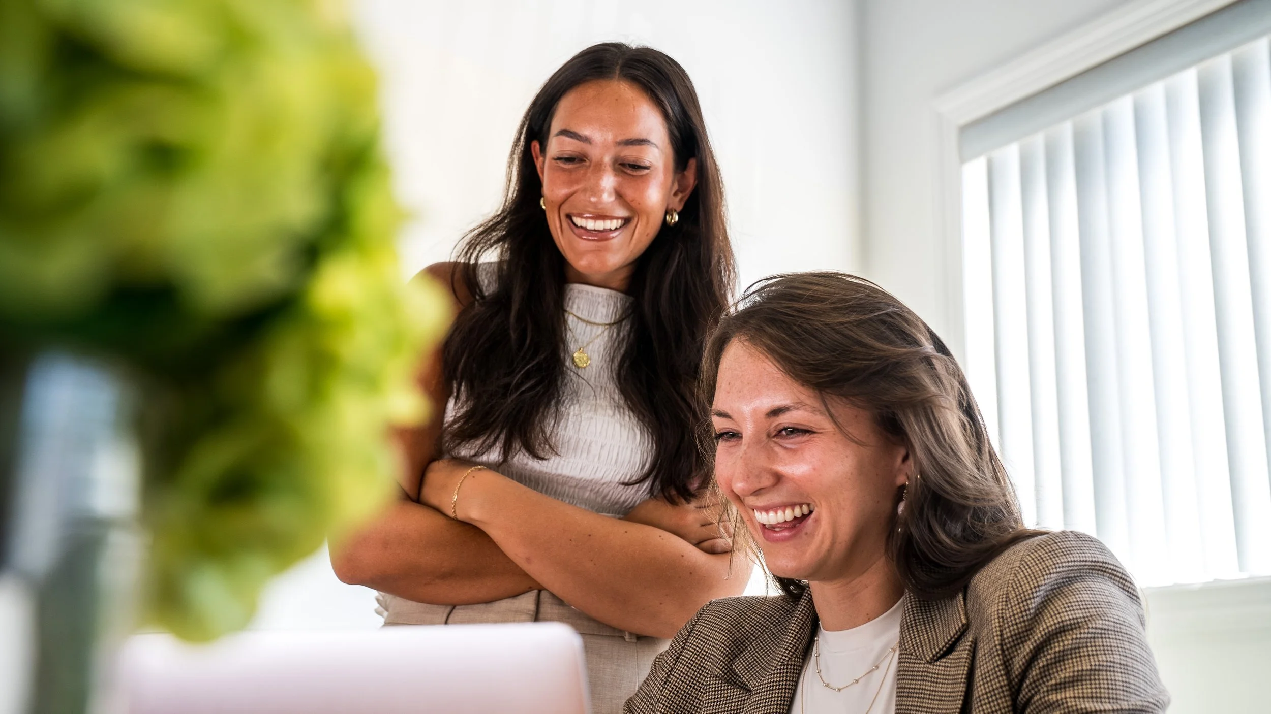 Haley Ursillo and Allison Dananzo  looking at a computer screen and smiling, with a potted plant in the foreground and window blinds in the background.