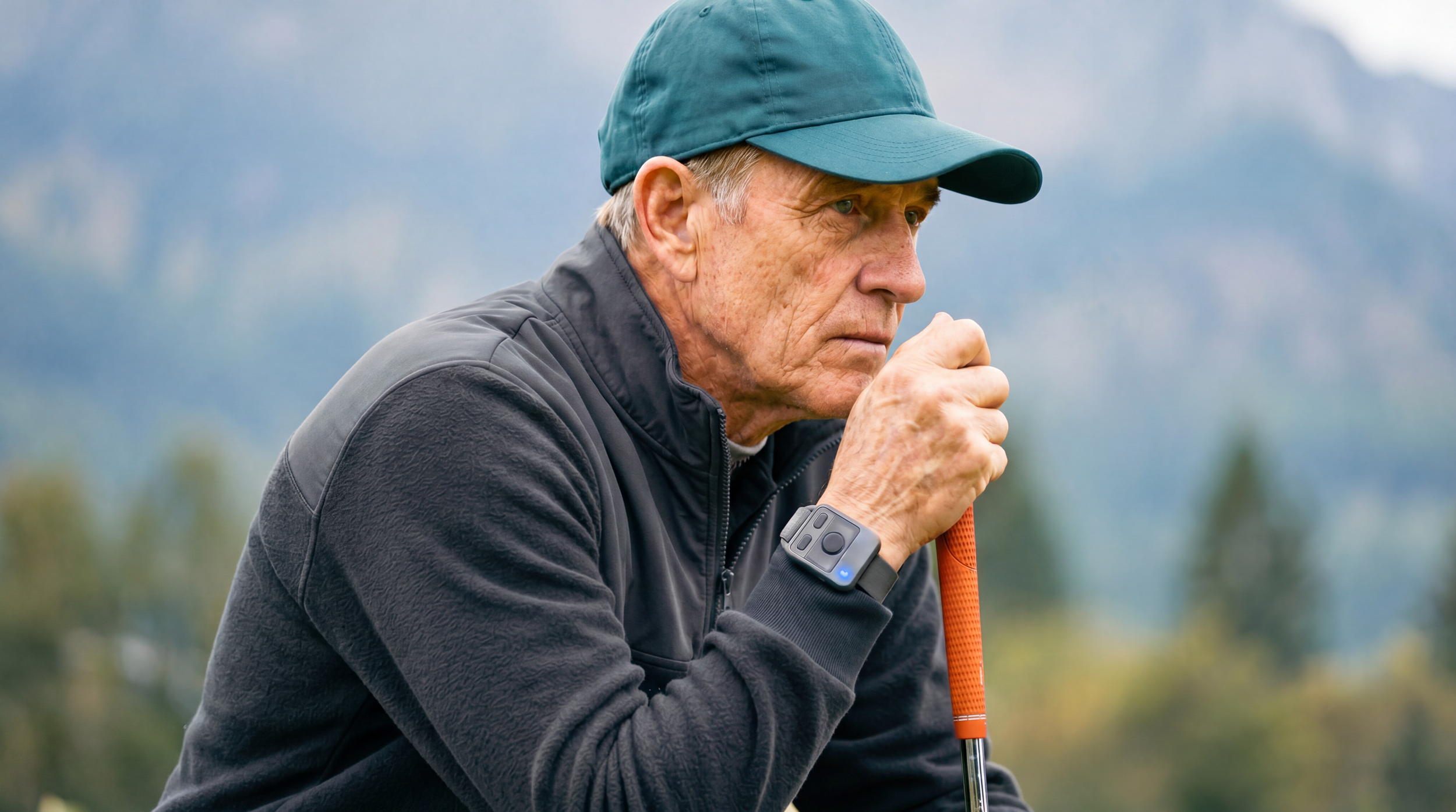 Older man in a teal baseball cap and gray outdoor jacket holding a golf club and looking pensively into the distance outdoors with a blurred mountain background.