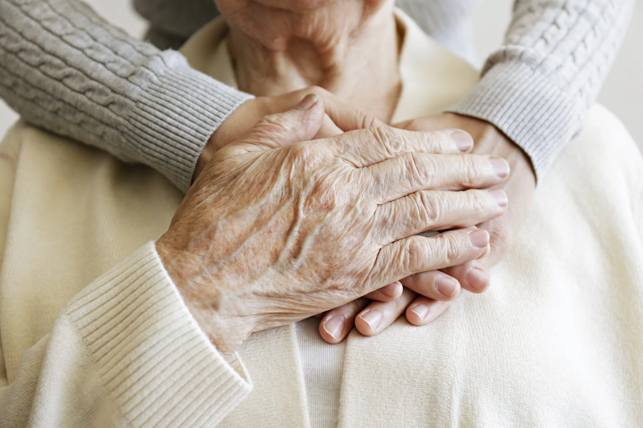 Close-up of elderly person with wrinkled hands resting on their chest, with a caregiver's hands gently holding theirs. The person is wearing a cream-colored sweater, and the caregiver is wearing a gray cable-knit sweater.