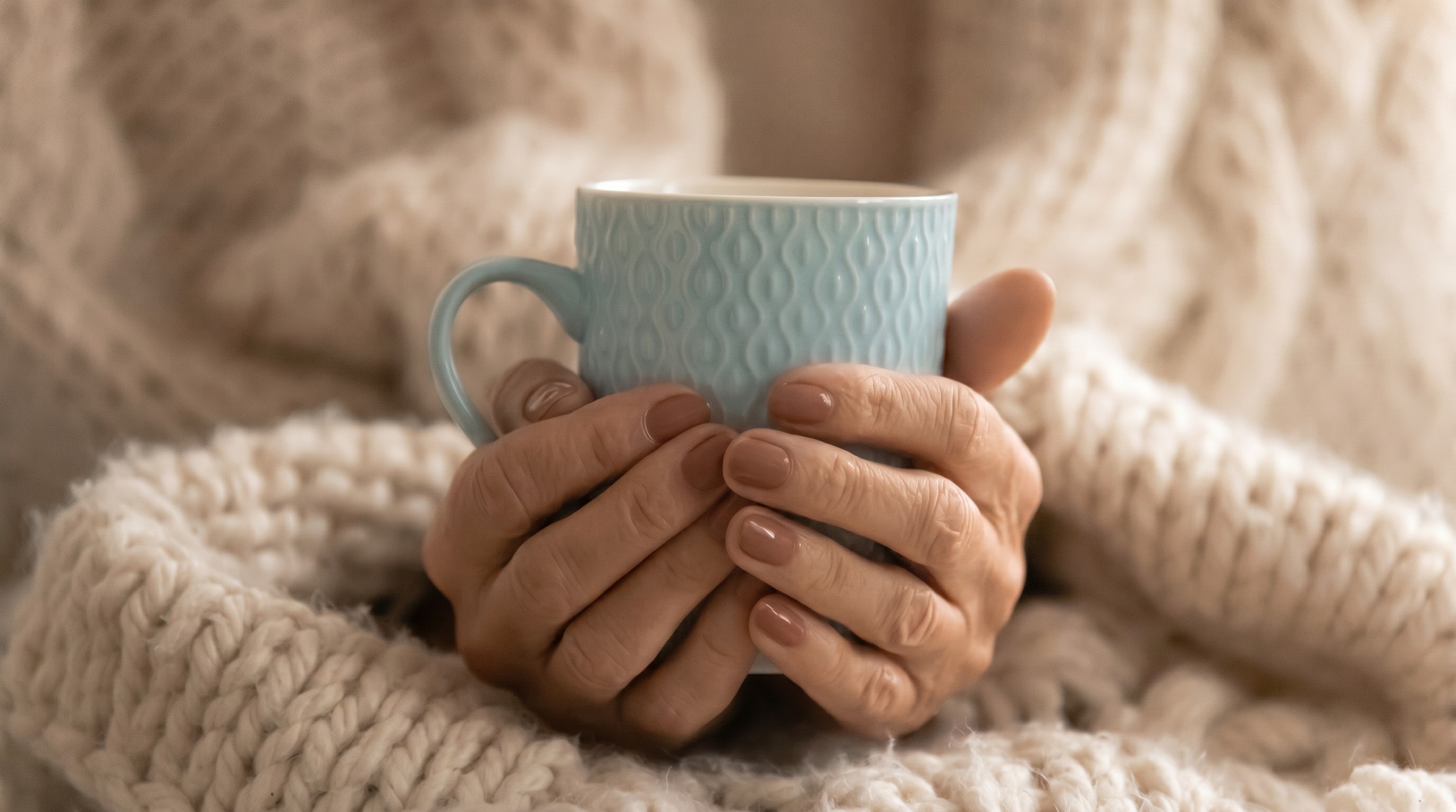 A person with well-manicured nails holding a light blue textured ceramic mug, dressed in a chunky cream-colored knit sweater.
