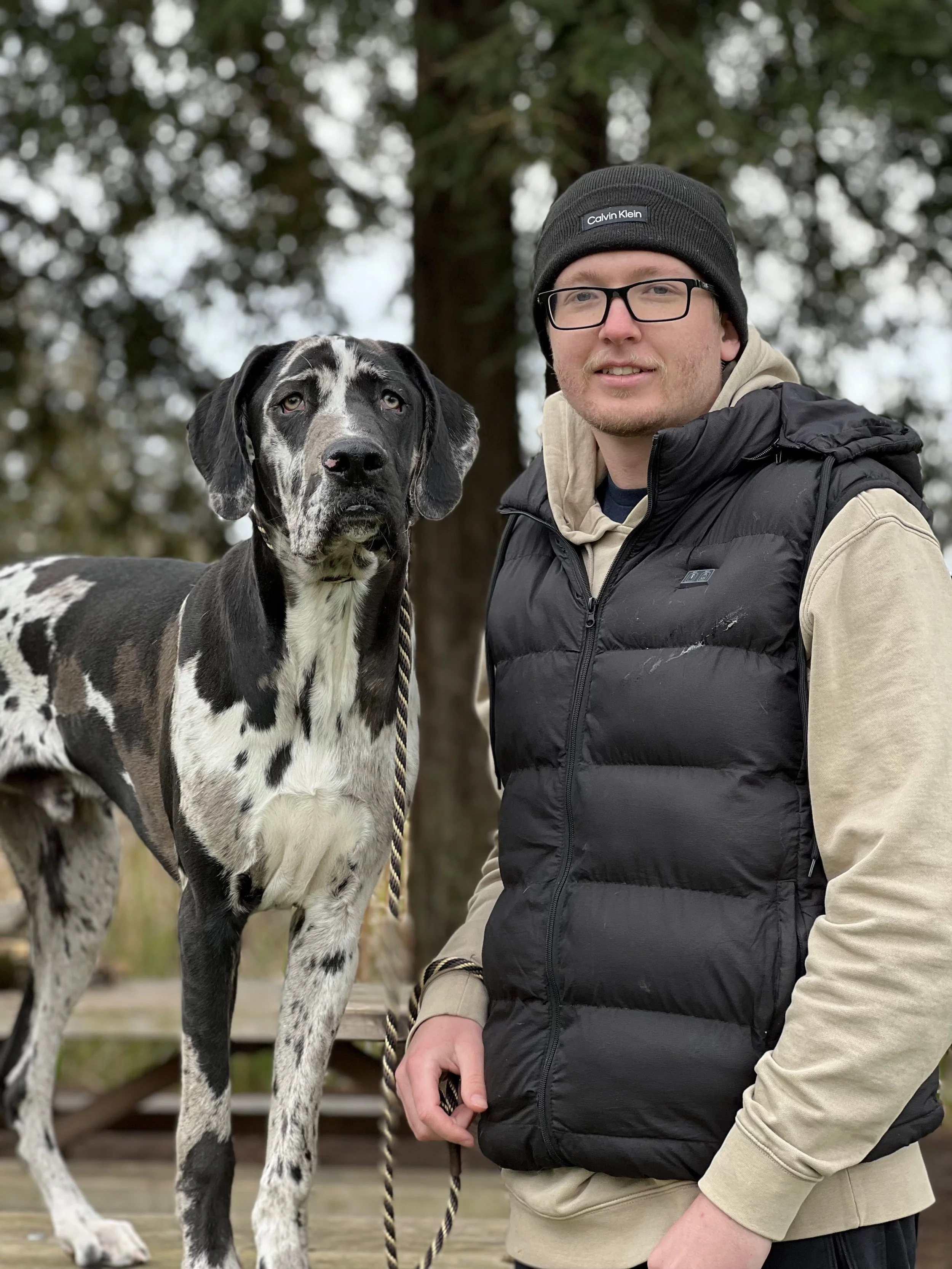 A man posing outdoors with a Great Dane dog on a leash, surrounded by trees.
