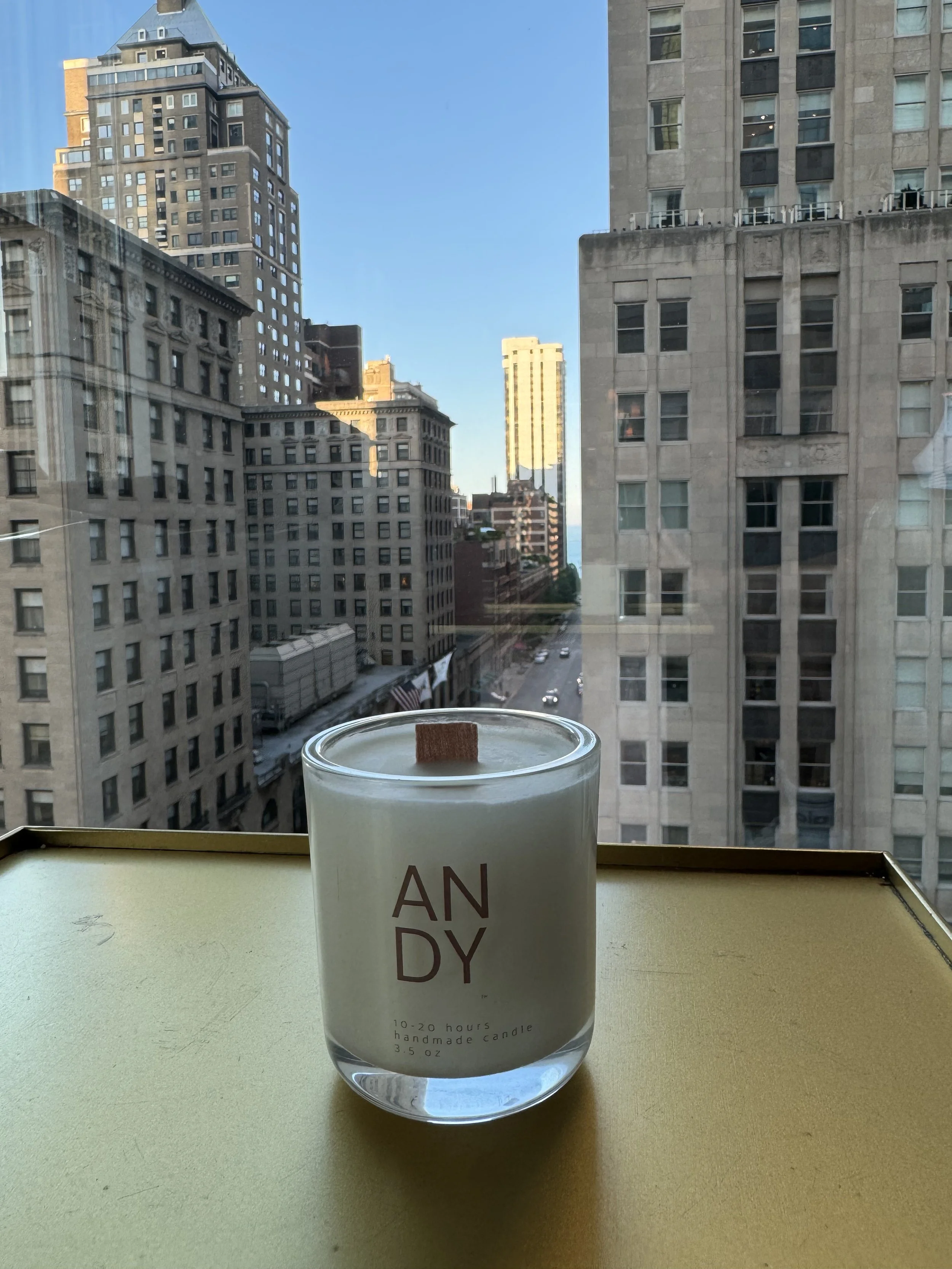 A white candle in a clear glass holder with minimal text on the front, sitting on a yellow surface with a cityscape of tall buildings and streets visible through a window in the background.
