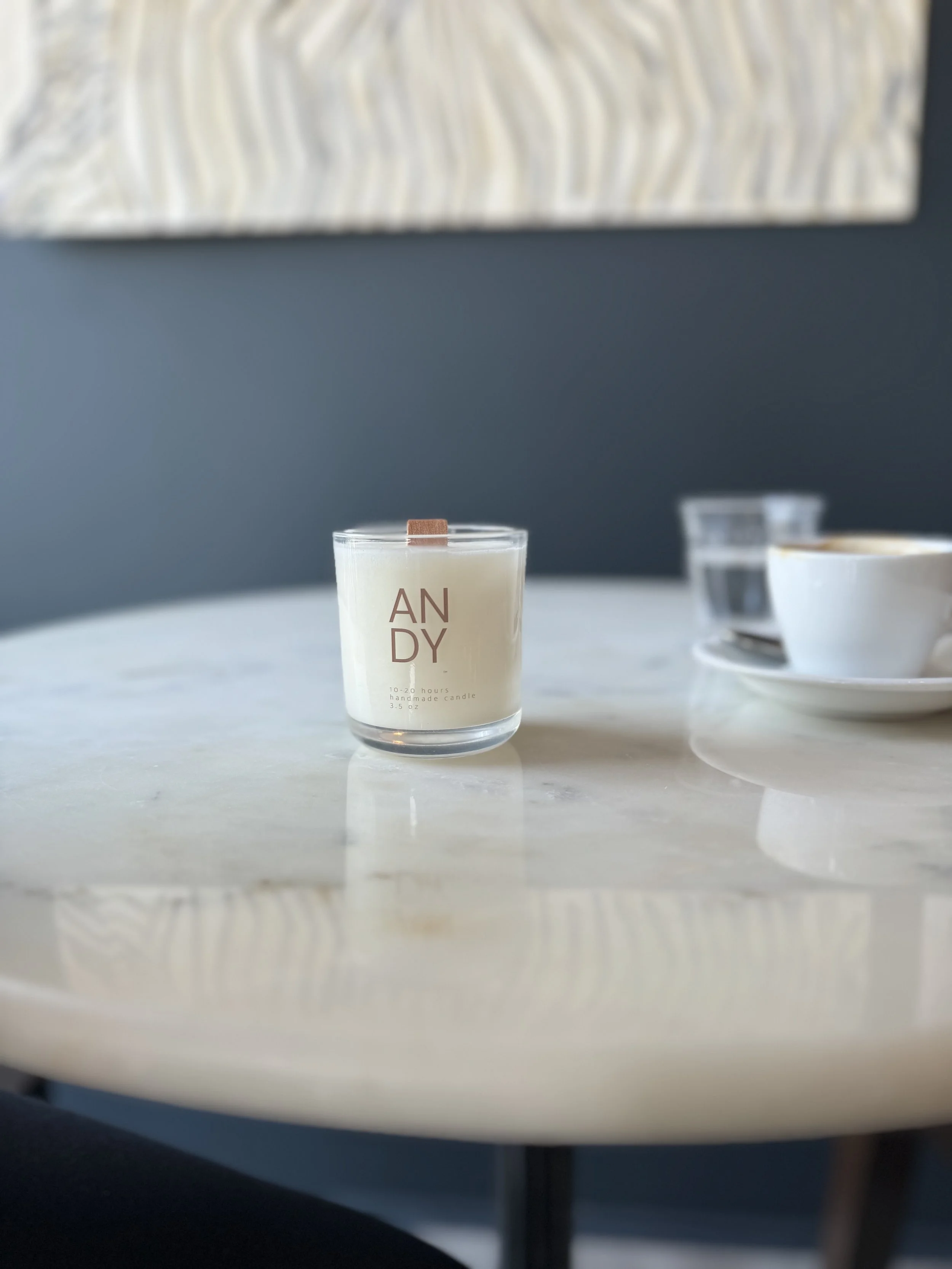 A white candle in a clear glass holder with the words 'ANDY' printed on it, placed on a white surface with a dark gray wall in the background. Part of a white teacup and a small glass container are also visible to the right.
