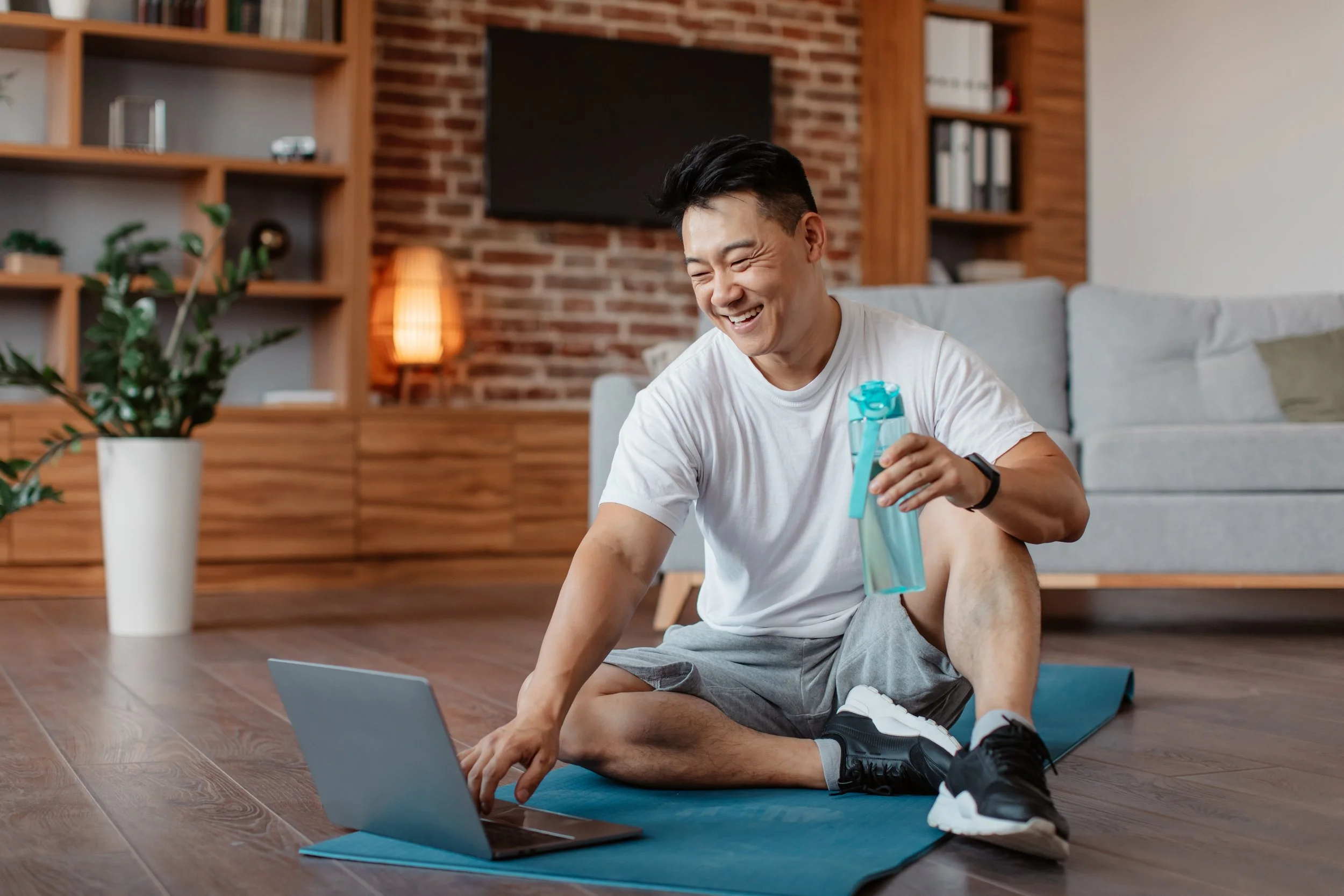 A man smiling and sitting on a yoga mat in a living room, holding a water bottle, with a laptop in front of him, and a large window, brick wall, shelves, and houseplant in the background.