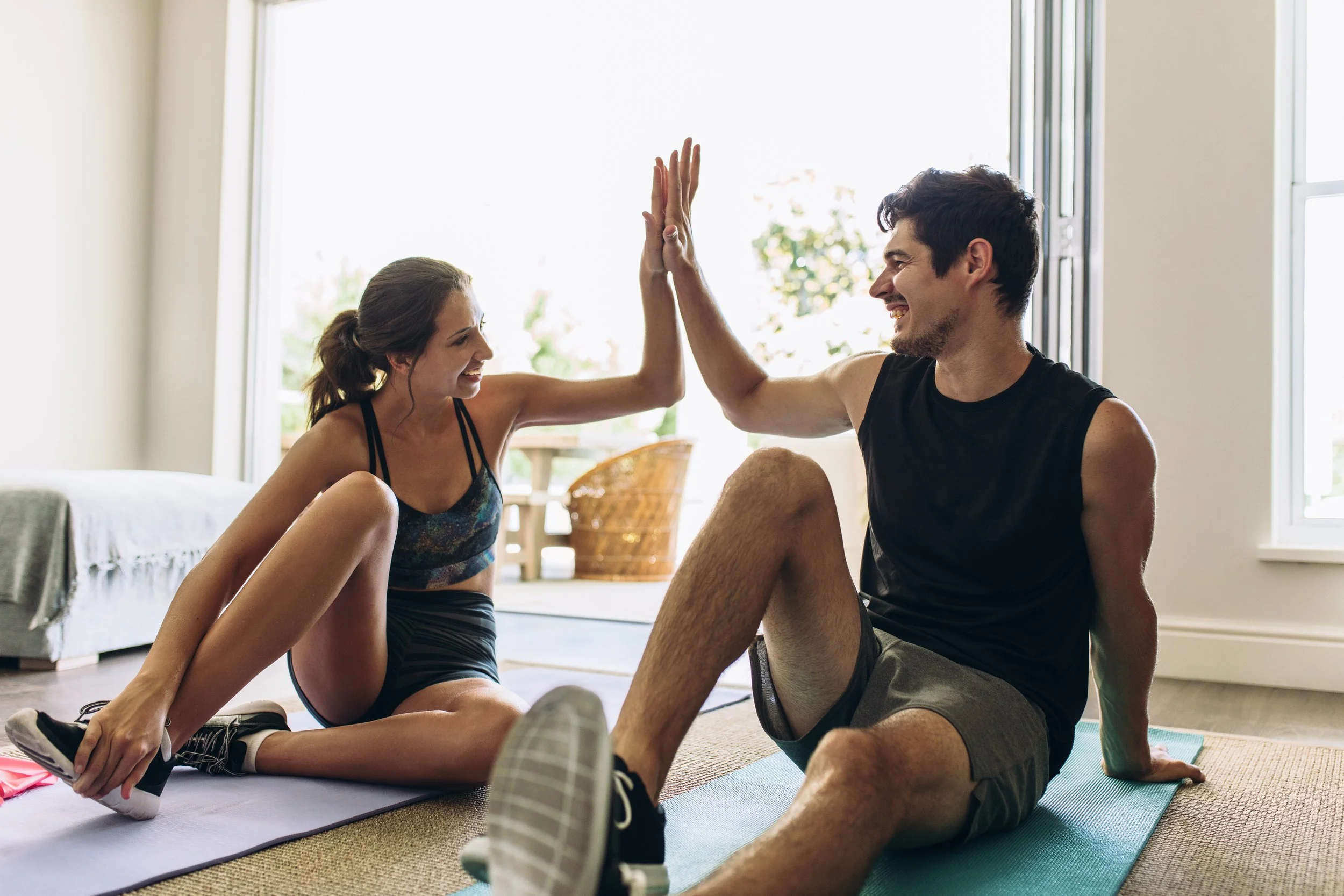 A young woman and a young man are sitting on the floor of a bright room, high-fiving each other during a workout session. They are dressed in athletic wear and smiling at each other. The room has large windows letting in natural light, with some furniture and plants visible in the background.