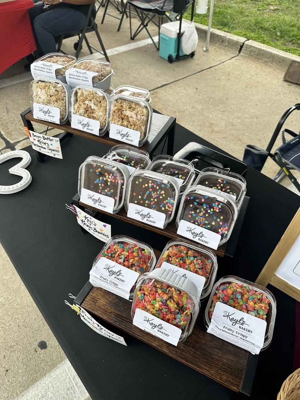 Display of assorted baked goods, including marshmallow squares, colorful brownie bites, and fruity cereal marshmallow squares, on a black table at an outdoor market.