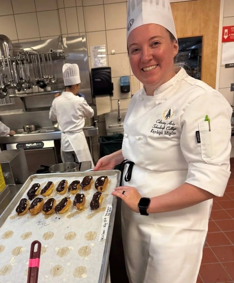 A smiling female chef in a white uniform and chef hat holding a tray of éclairs topped with chocolate in a professional kitchen.