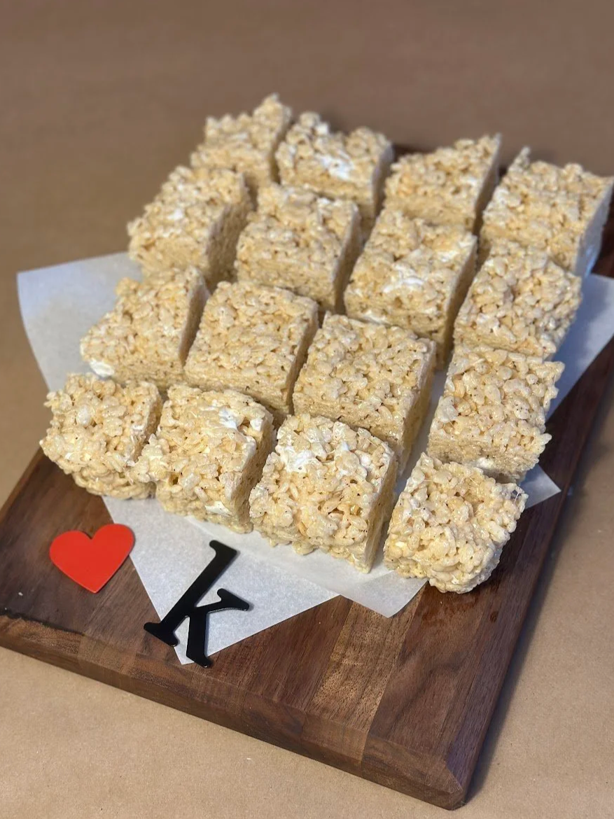 A wooden serving board with chopped rice crispy treats topped with marshmallows, arranged in rows. The treats are square, coated with rice cereals, and the board has decorative red and black letters and a red heart.