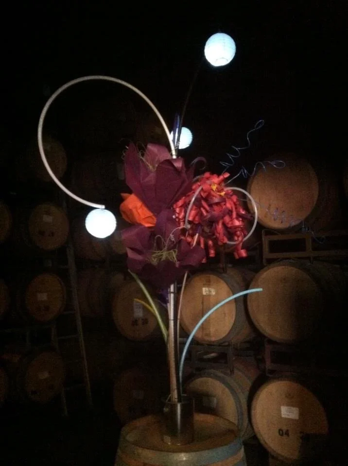 Decorative floral arrangement with purple and red flowers, illuminated by small globe lights in a dark setting with wooden barrels in the background.