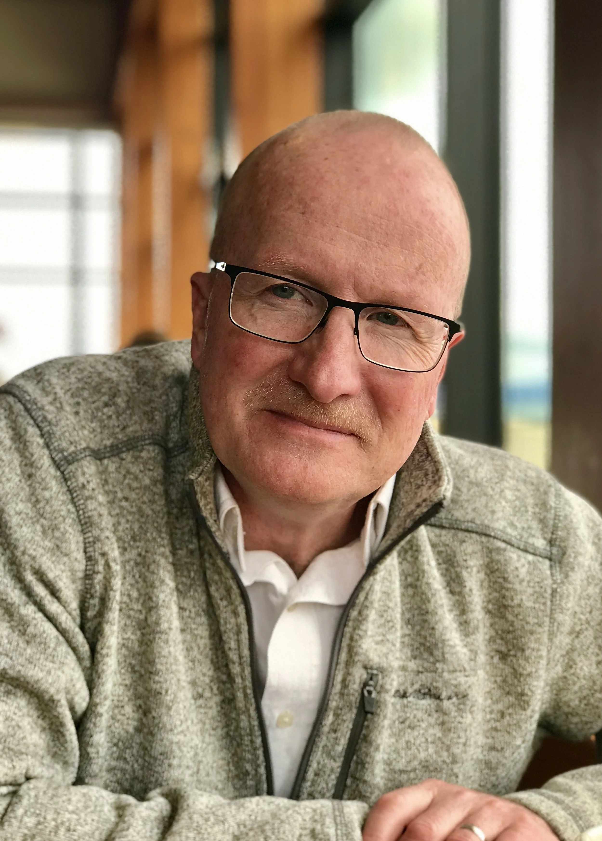 A close-up photo of a smiling elderly man with glasses, wearing a light grey fleece jacket over a white shirt, sitting indoors near a window.